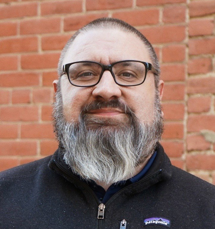 Portrait of a smiling man with glasses, a beard, and short hair, standing outdoors in front of a brick wall.