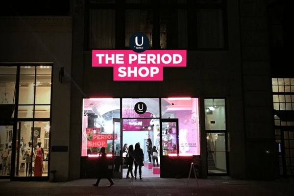 The storefront of 'The Period Shop' with bright pink signage and silhouettes of people inside and outside at night.