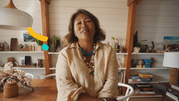 A woman with shoulder-length brown hair wearing a beige shirt and layered necklaces sitting at a wooden table in a cozy room with wooden beams, white walls, and bookshelves.