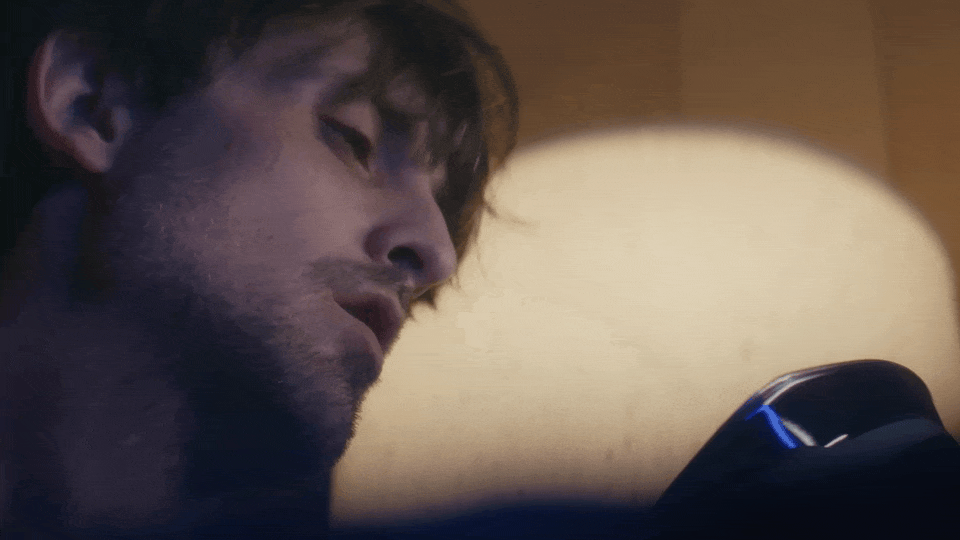 Close-up of a young man with dark hair and stubble, lying with his eyes closed, illuminated by the screen of a device in a dimly lit room.