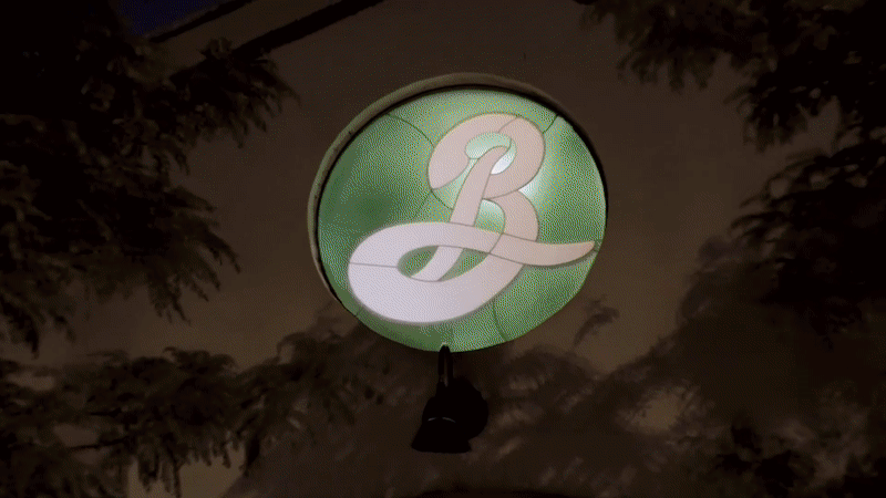 Illuminated circular sign with a stylized letter 'B' in white against a green background, hanging outdoors at night surrounded by trees.