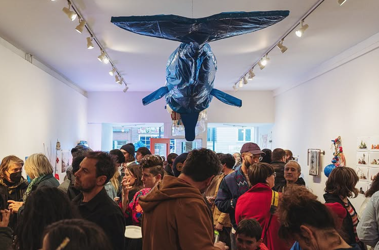 Crowd of people attending an art gallery or exhibition with artwork and sculptures displayed on the walls, and a large art installation hanging from the ceiling resembling a whale.