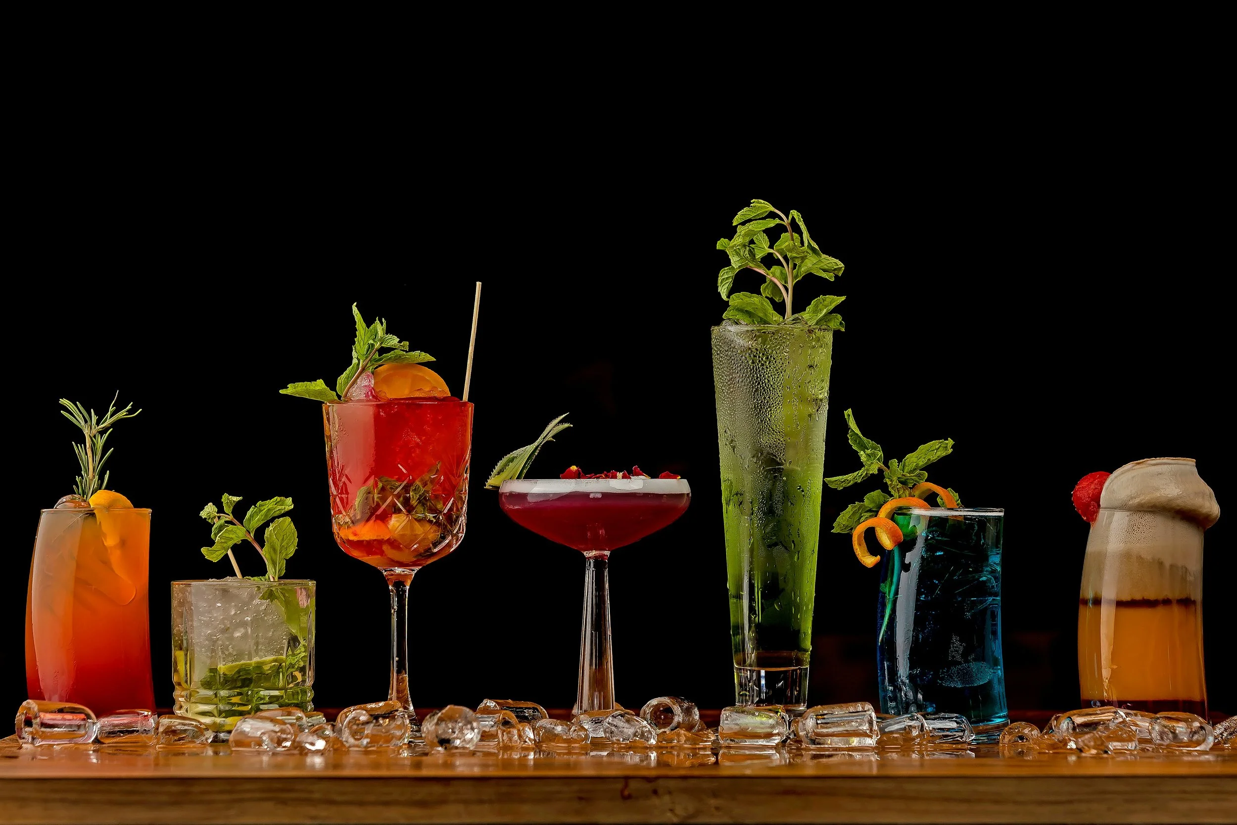 A variety of colorful cocktails and drinks served in different glasses, garnished with fruits, herbs, and decorative elements, placed on a wooden surface with ice cubes, against a black background.