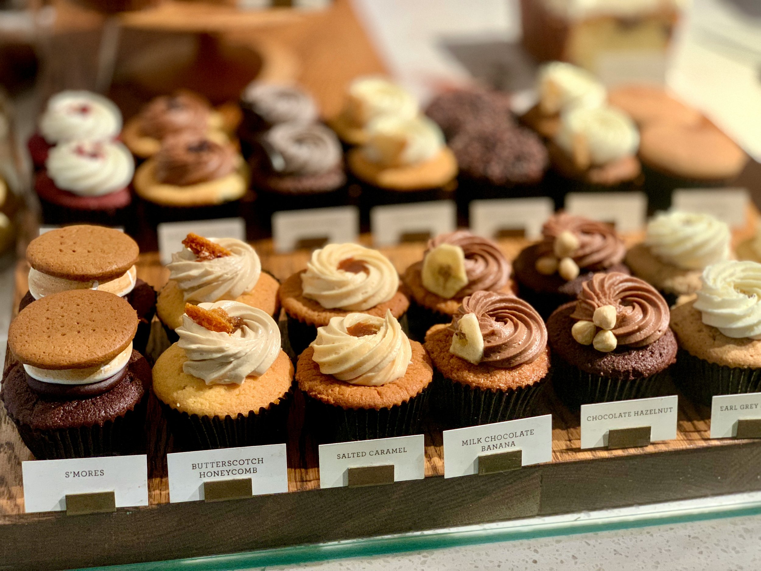 Assorted cupcakes with various frosting toppings on a tray at a bakery, including s'mores, salted caramel honeycomb, milk chocolate banana, chocolate hazelnut, and Earl Grey.