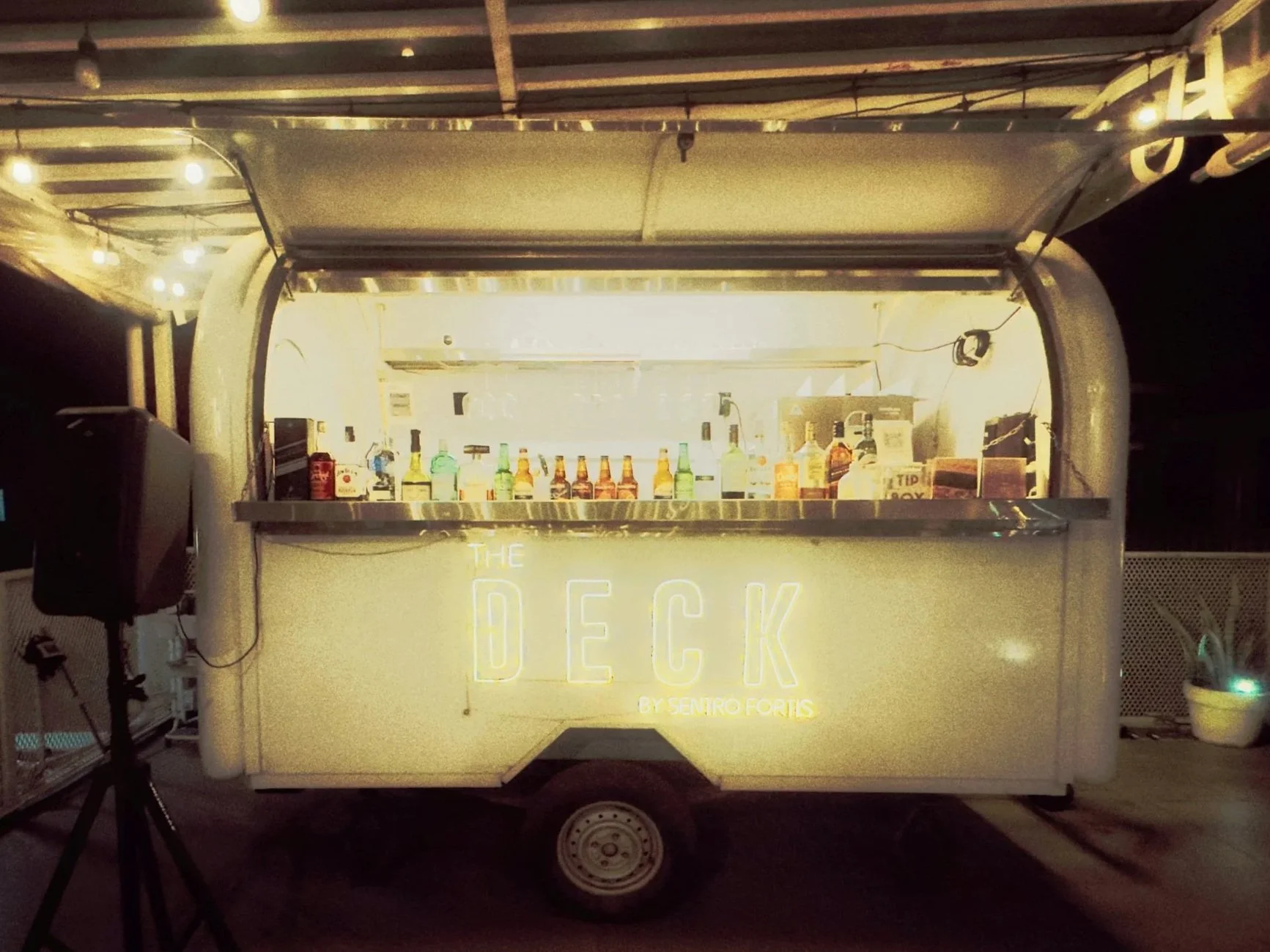 A mobile bar cart named 'The Deck' by Sentro Fortis, illuminated with neon lights, displaying various bottles of alcohol and mixers inside an open trailer at night.