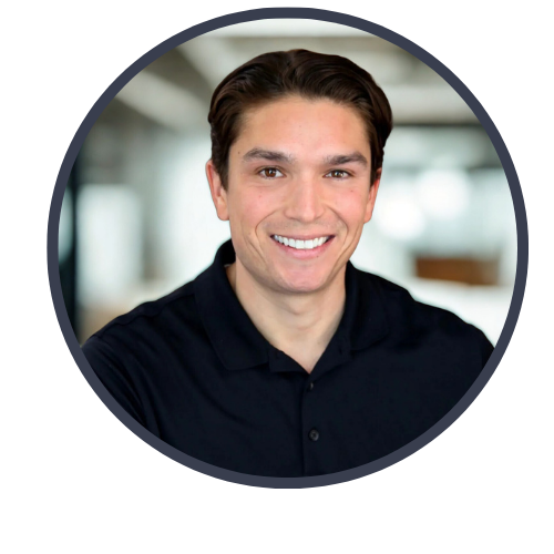 A smiling young man with brown hair wearing a black collared shirt, indoors with a blurred background.