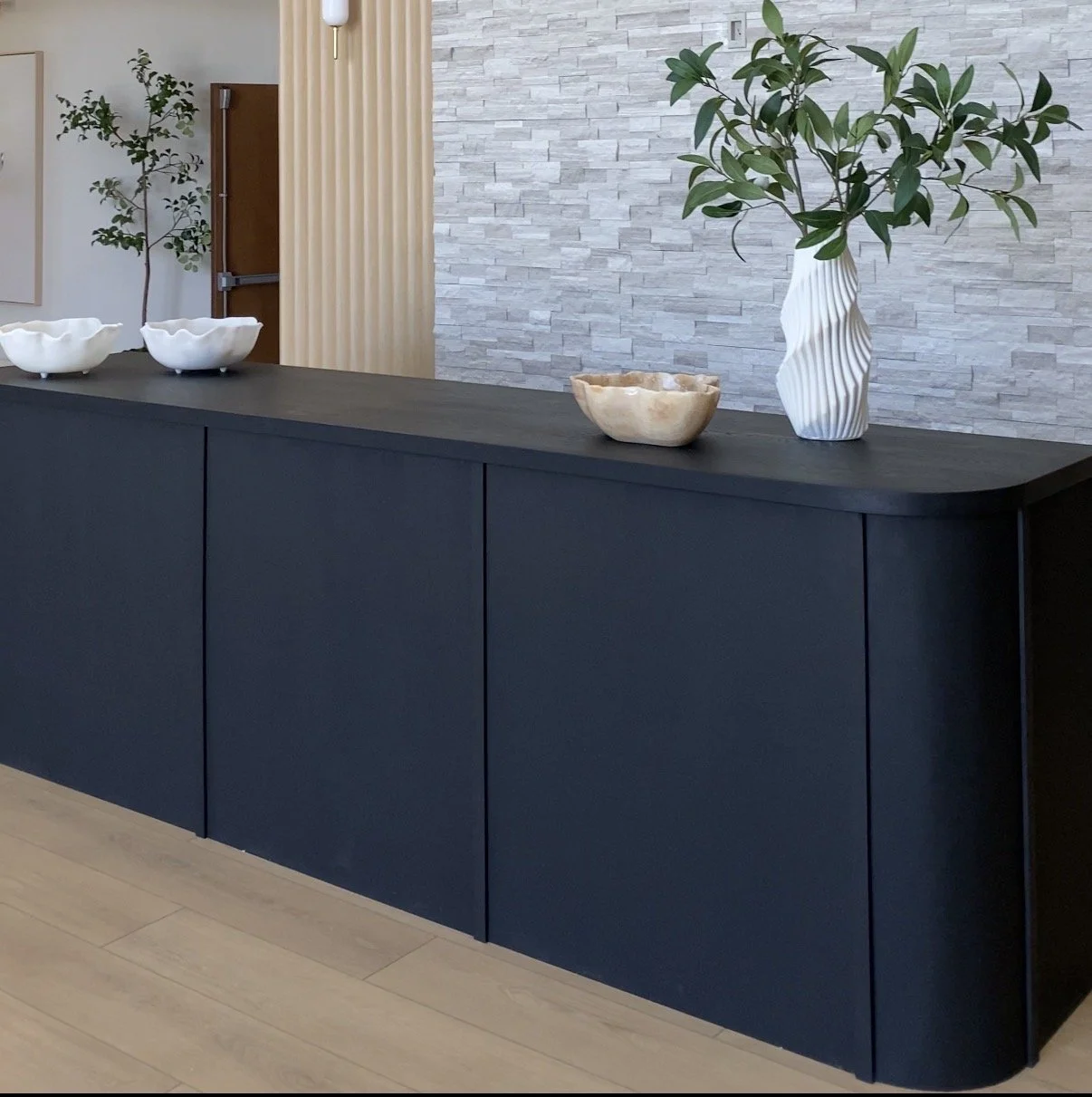 A black sideboard with decorative bowls and vases, along with a tall vase holding green leafy branches, positioned against a textured light gray wall in a modern interior space.