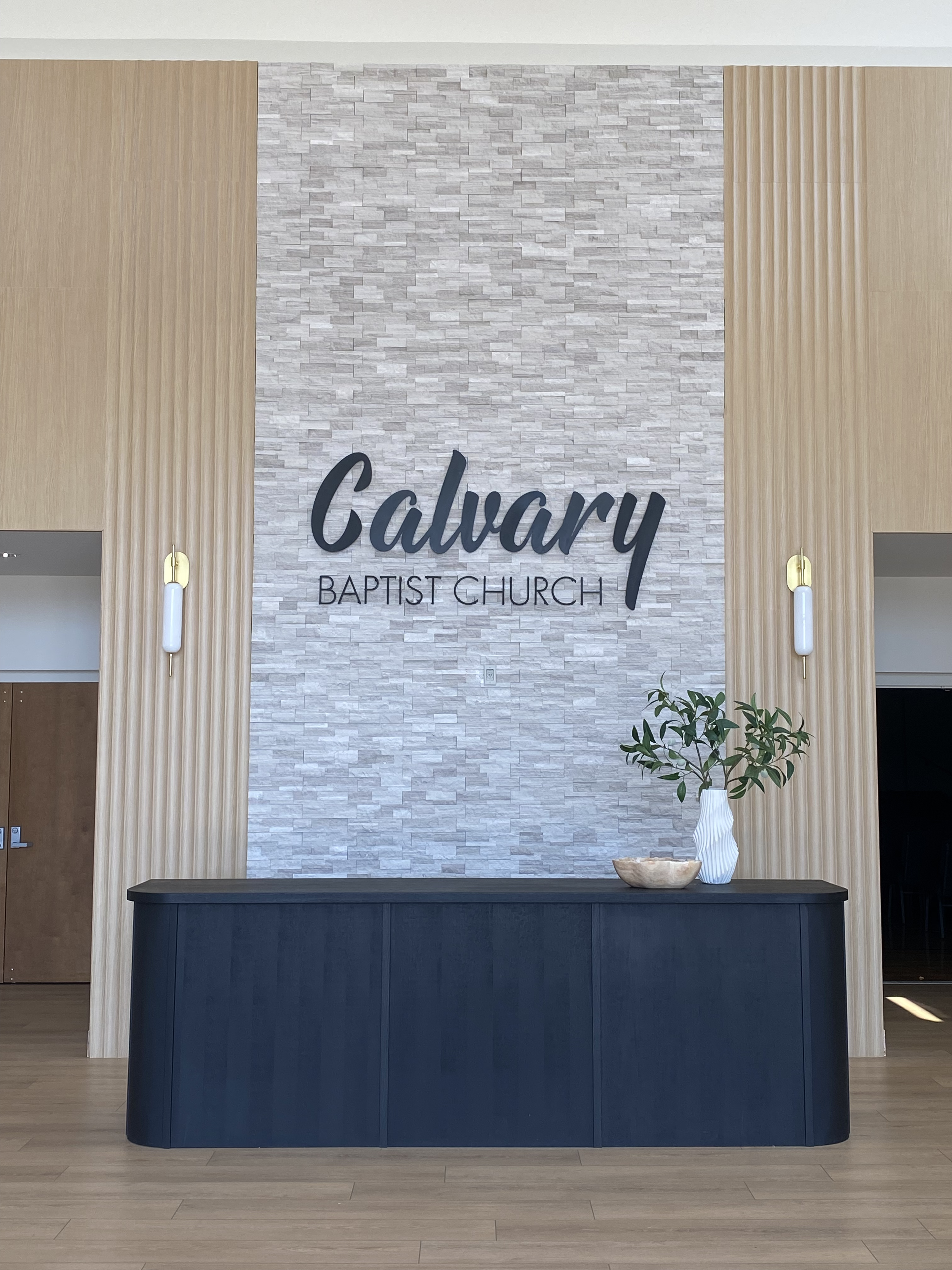 Interior view of Calvary Baptist Church's reception area with a black reception desk, a white vase with greenery, a wooden bowl, and a textured stone wall featuring the church's name in large black letters. built by B&H design & Build