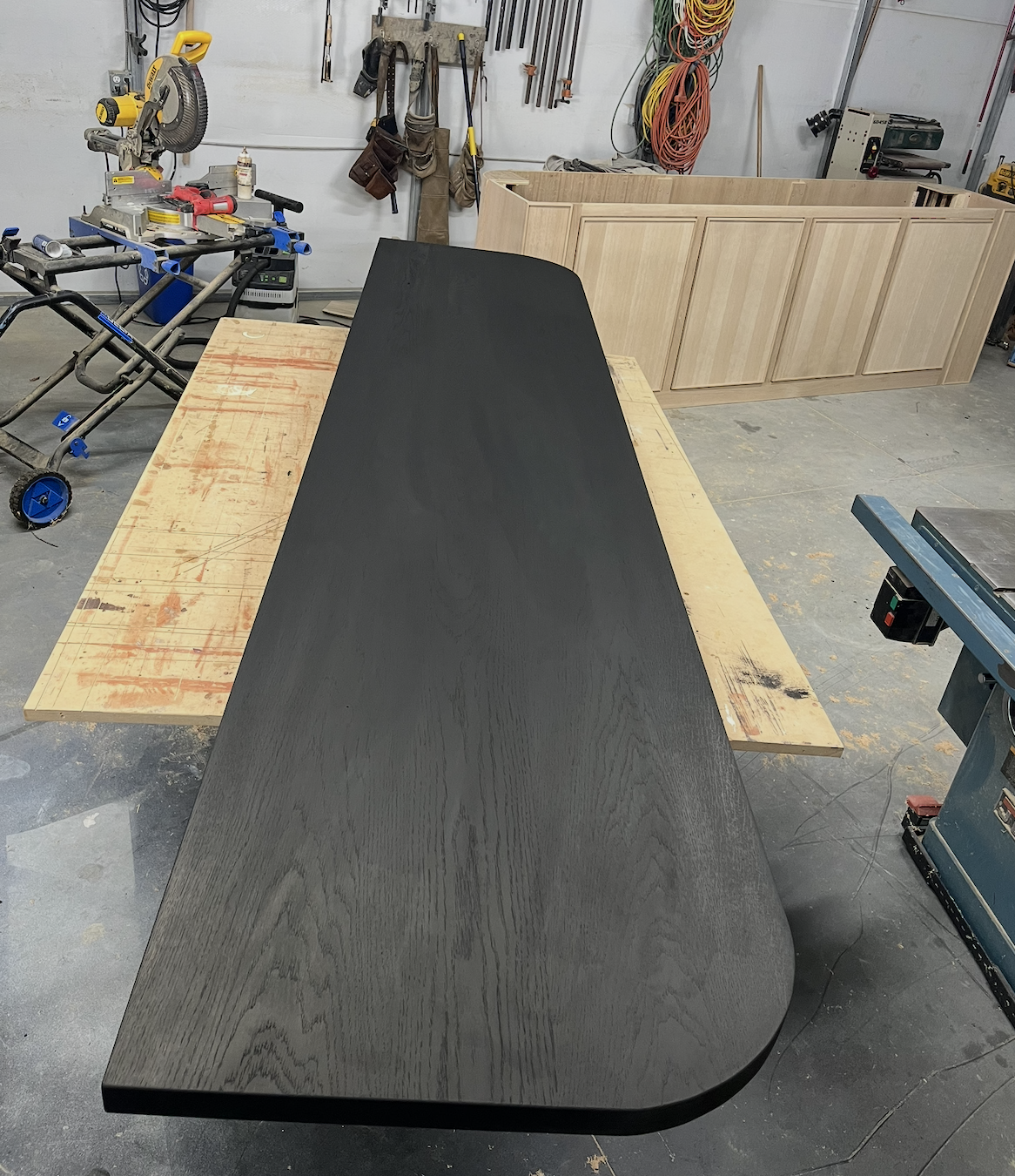 A woodworking workshop with a long, dark wood table in the foreground, a workbench with tools and clamps, and a partially assembled wooden cabinet in the background.