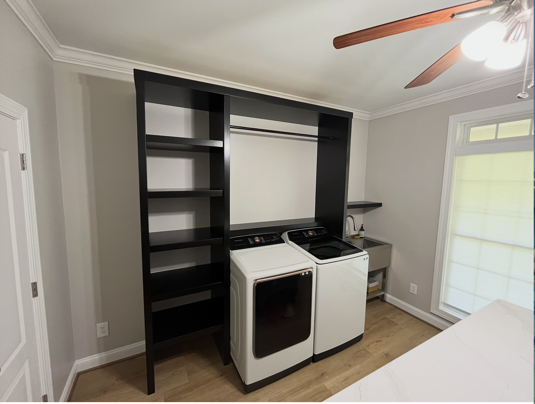 Laundry room with black shelving unit, washing machine, dryer, stainless steel sink, small shelf, and window with blinds.