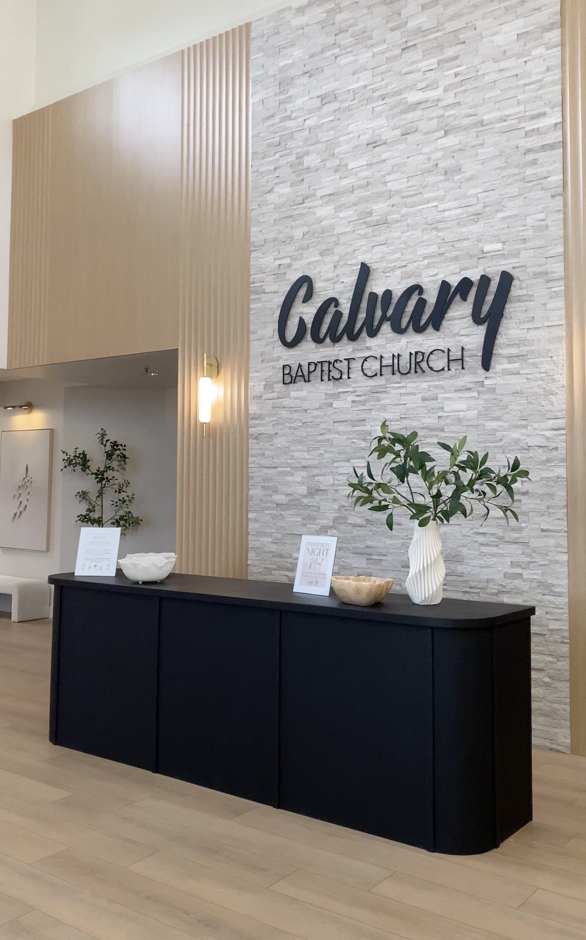 Interior of Calgary Baptist Church with a black console table holding decorative bowls and a vase with greenery, against a stone accent wall with the church's name in large black letters.