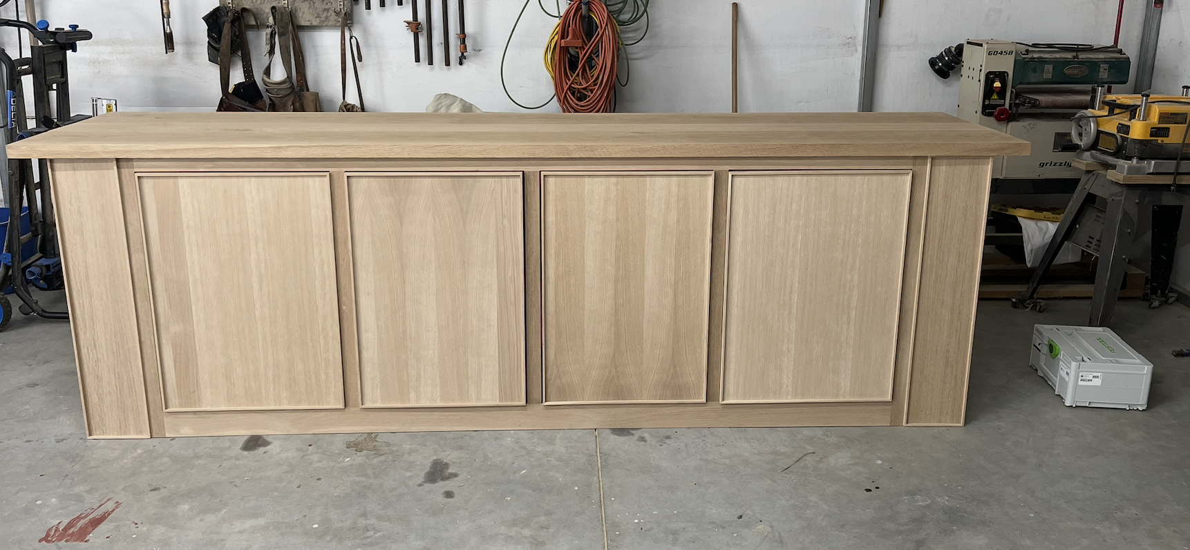 A wooden cabinet or sideboard in a workshop, with four paneled doors, made of light-colored wood, on a concrete floor.