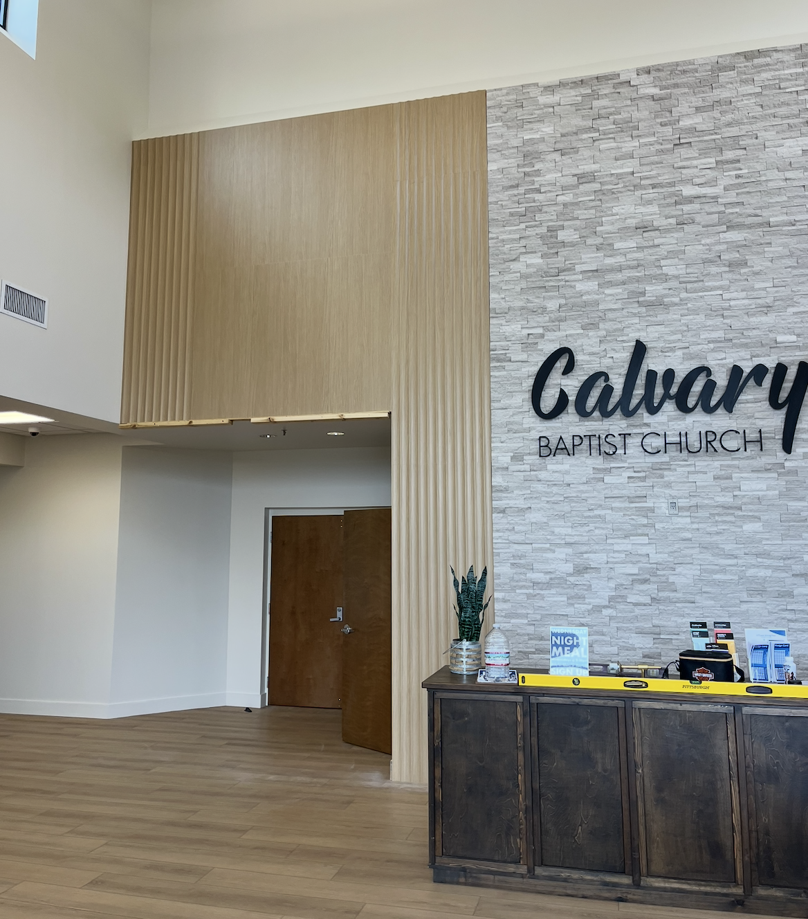 Inside Calvary Baptist Church with a wooden front desk, potted plant, and signage on a brick wall.