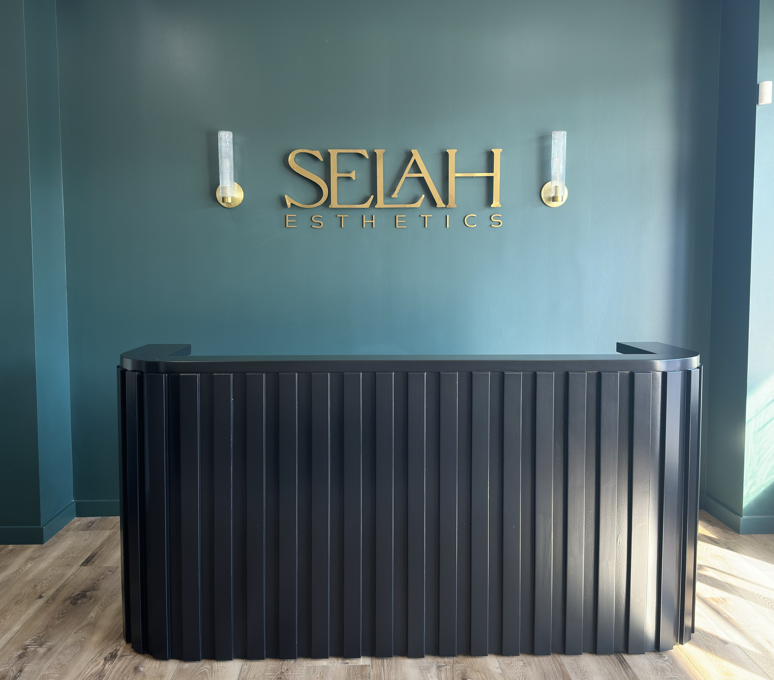 Reception desk in front of a teal wall with gold signage that reads 'SELAH ESTHETICS' and two modern wall sconces.