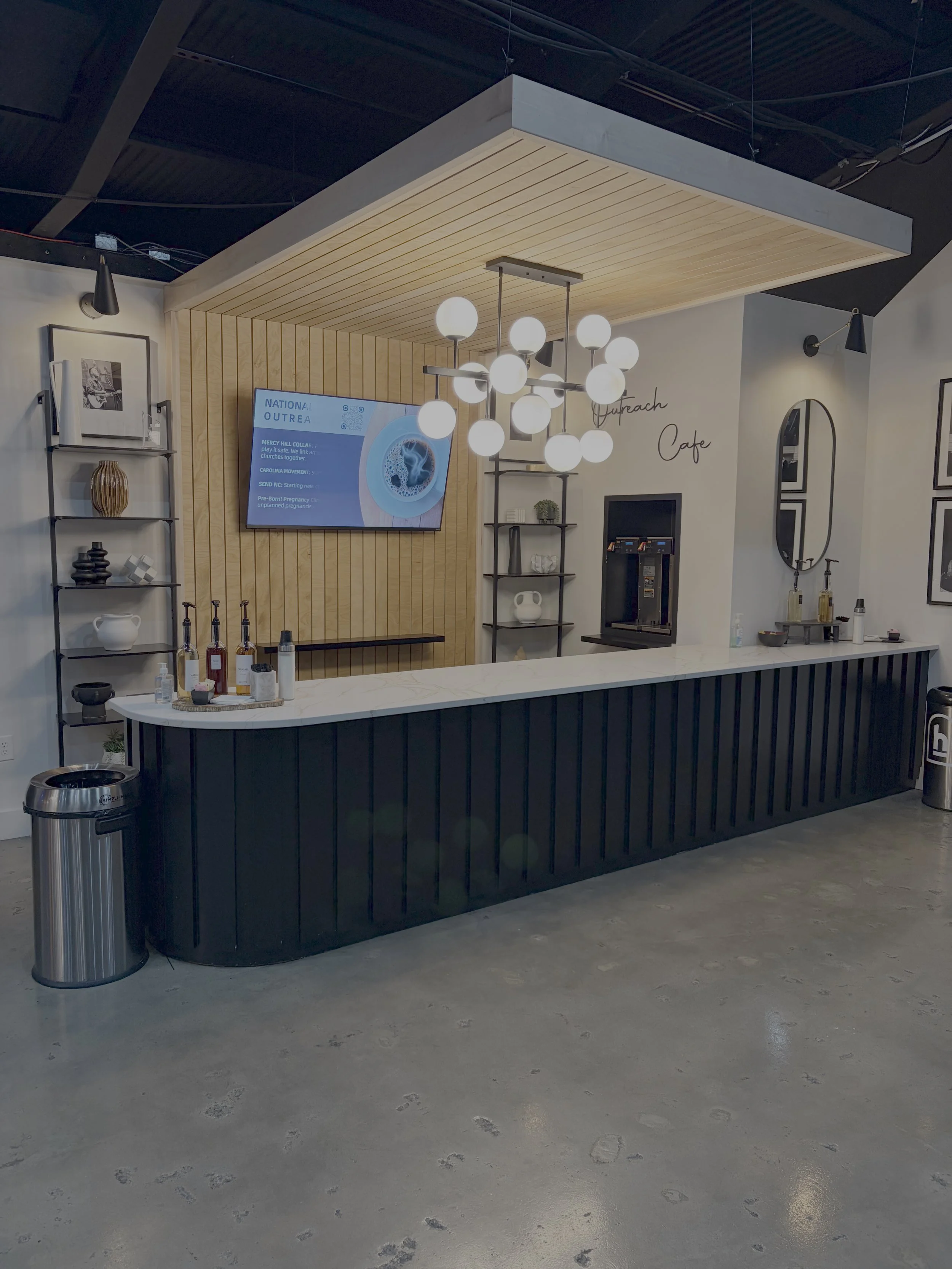 Modern cafe counter with a black base and white marble top, black shelving on white walls, a large chandelier with spherical bulbs, a digital screen, and a coffee machine, with minimalist decor and light wood accents.