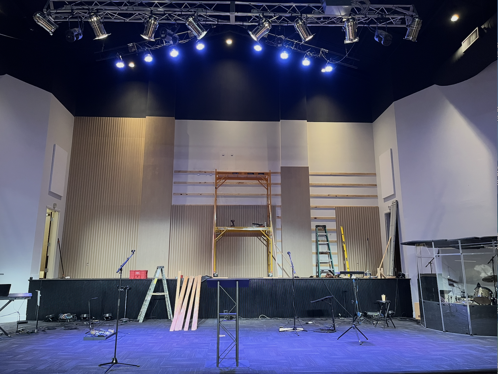Stage under construction with scaffolding, ladders, and various tools, lights overhead, and musical equipment on the floor.