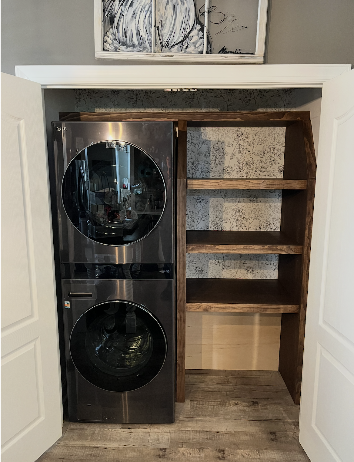A laundry closet with a stacked LG washer and dryer on the left and empty wooden shelves on the right, with floral wallpaper on the back wall.