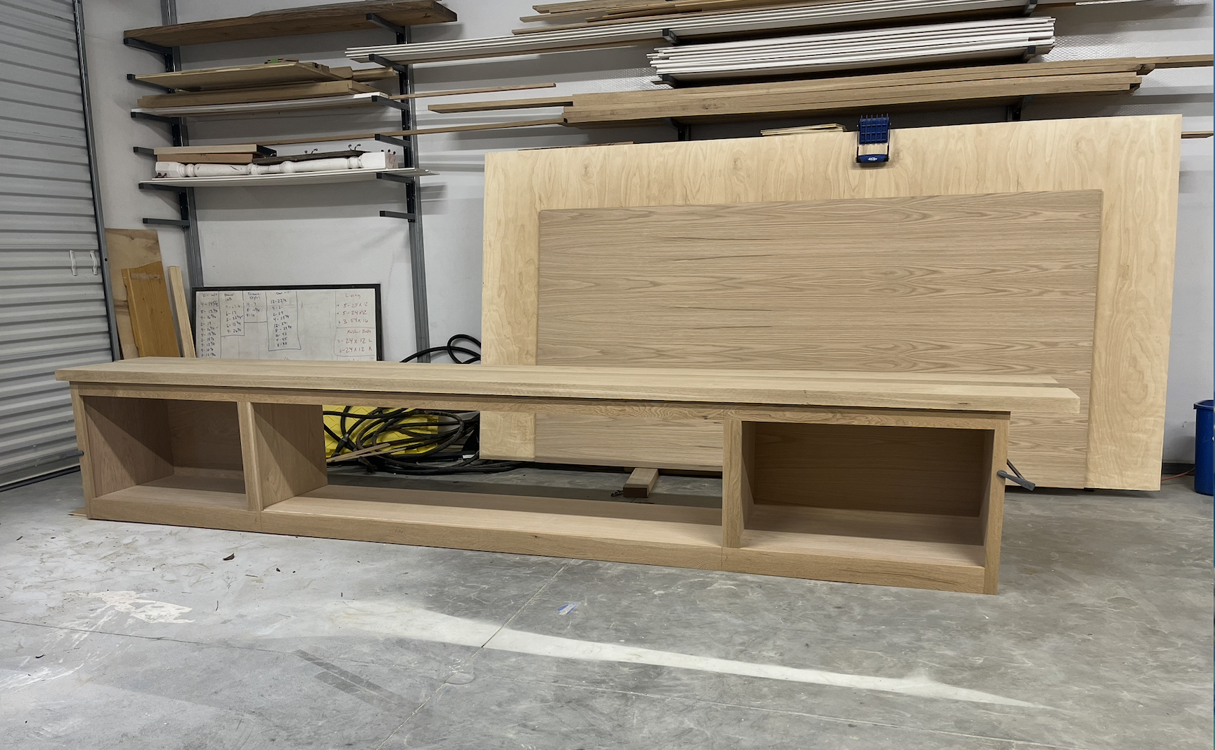 Unfinished wooden furniture piece in a workshop, with a large whiteboard showing measurements and notes, and shelves with wood planks in the background.