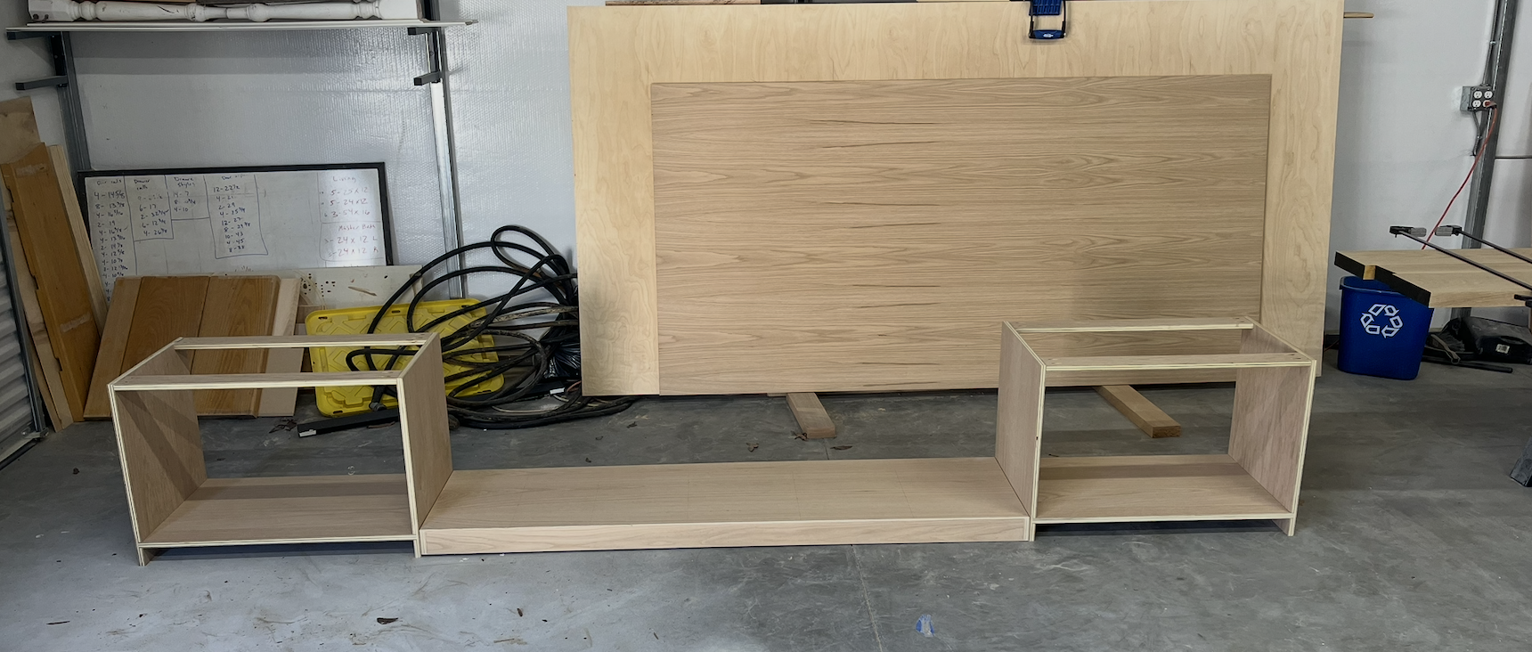Unassembled wooden furniture on a concrete garage floor, including two open-frame cube units and a long rectangular piece, with various woodworking tools and materials in the background.