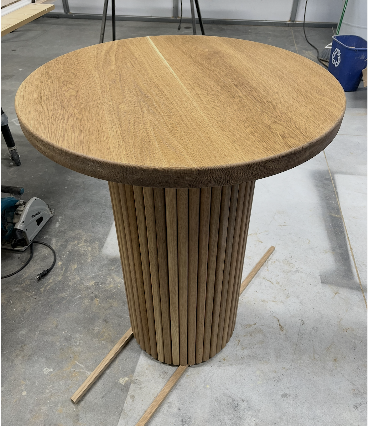 A custom wooden round table with a cylindrical base made of vertical wooden slats, in a workshop setting.