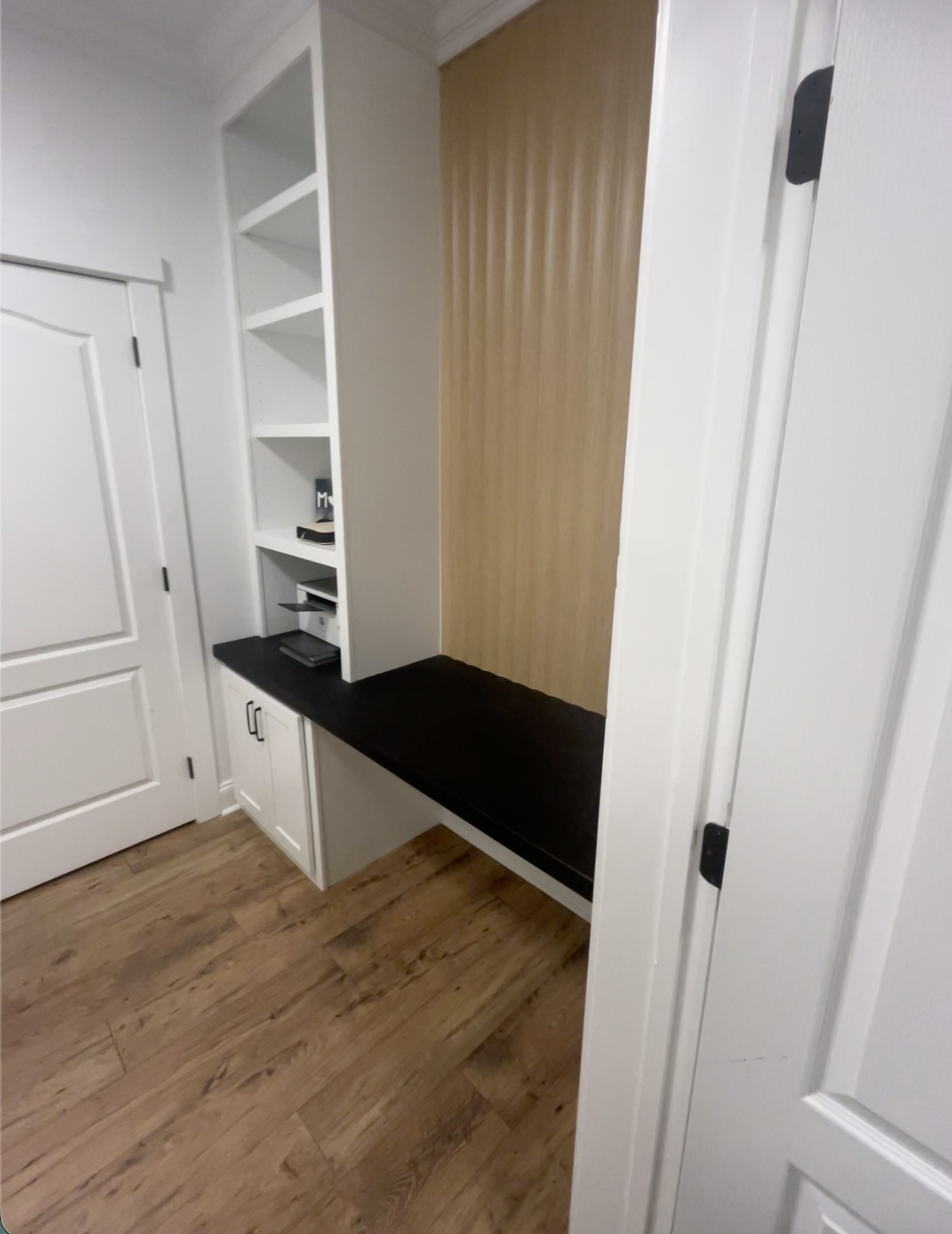 Built-in white shelving unit with black countertop, small cabinets below, and a wooden accent wall in a room with hardwood floors.