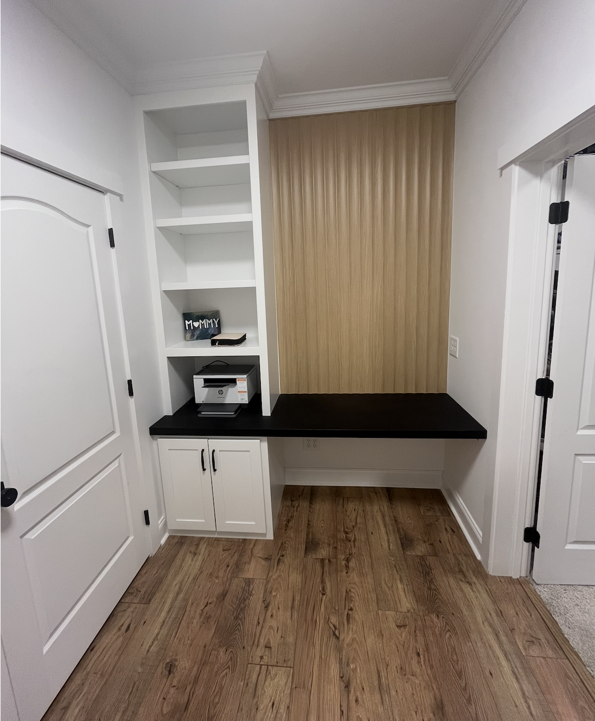Empty built-in desk and shelf unit with a black countertop, white cabinets, and a wooden accent wall in a room with wood flooring.