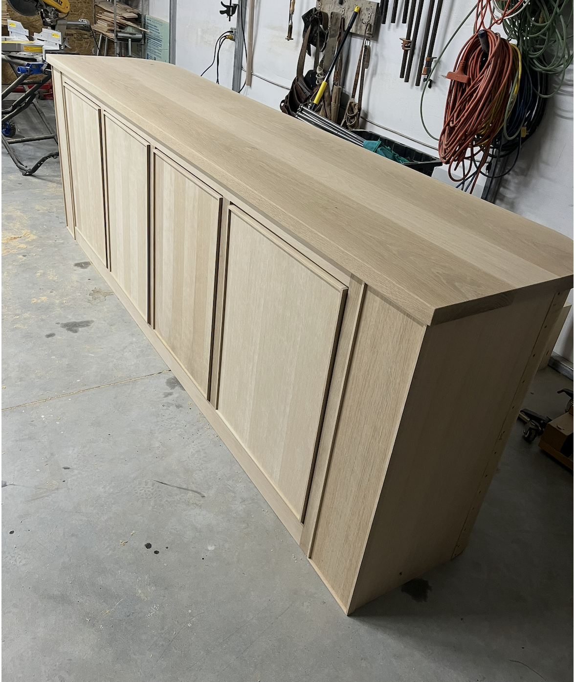A wooden cabinet with four doors on a workshop floor, surrounded by tools and hanging cords.
