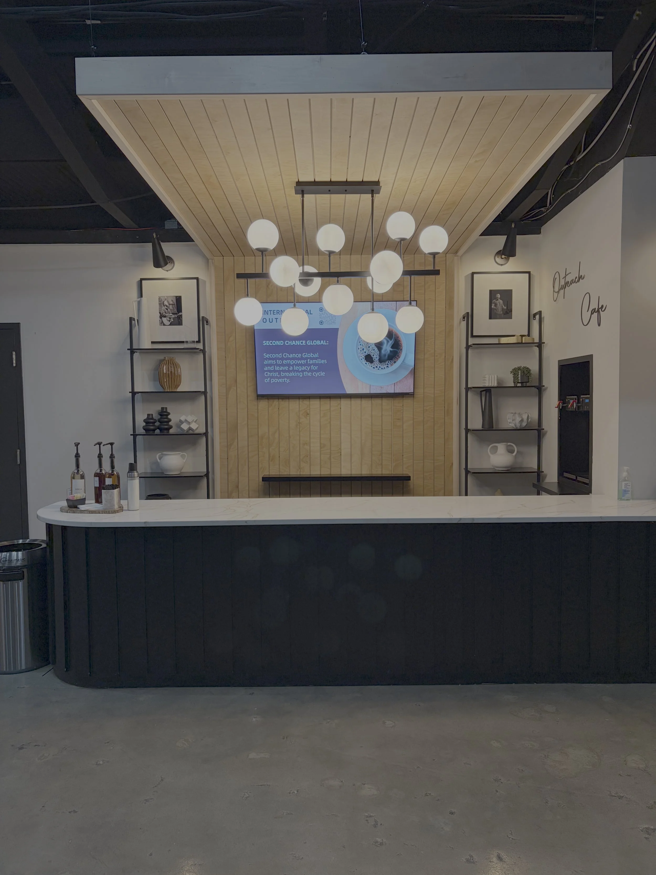 Interior of a modern café with a black and white color scheme, a white marble countertop, black shelving with decorative items, a flat screen display on a wooden wall, and modern lighting fixtures hanging from a wooden ceiling.