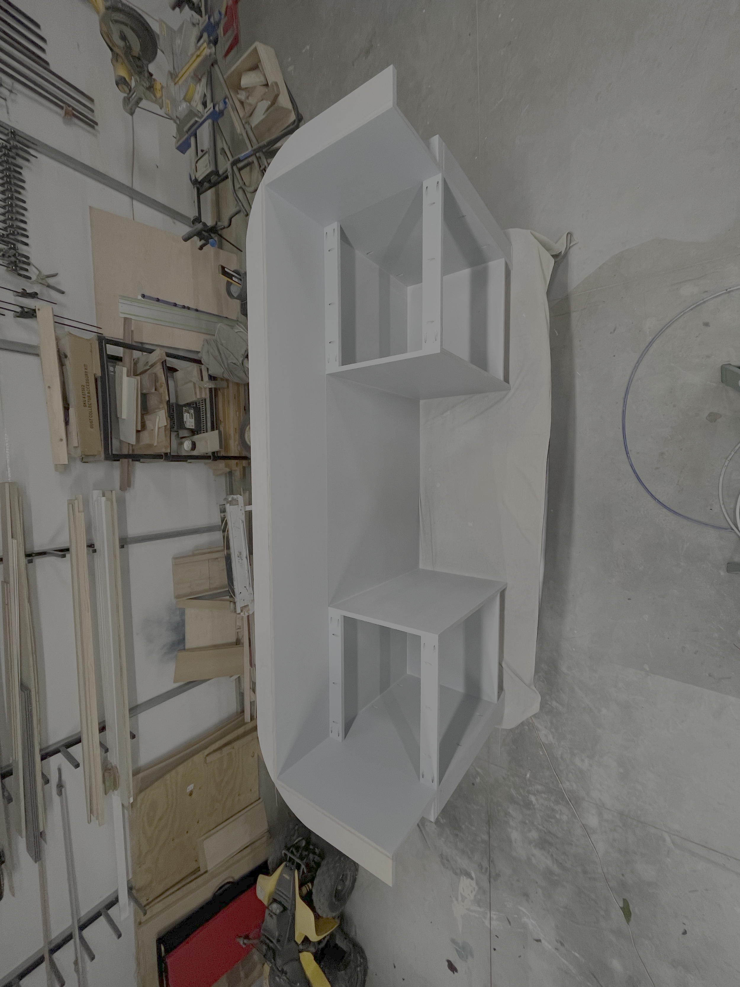White wooden table with two open shelves, surrounded by woodworking tools and equipment in a workshop.