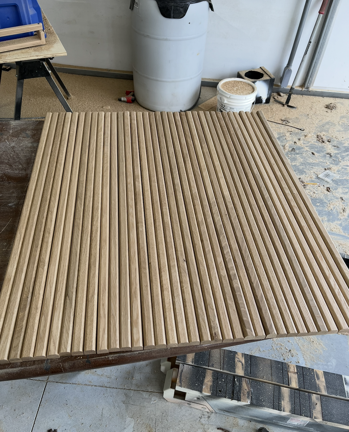 A workspace with a wooden slatted table, a large white trash can, a bucket of sand or wood stain, and woodworking tools and materials.