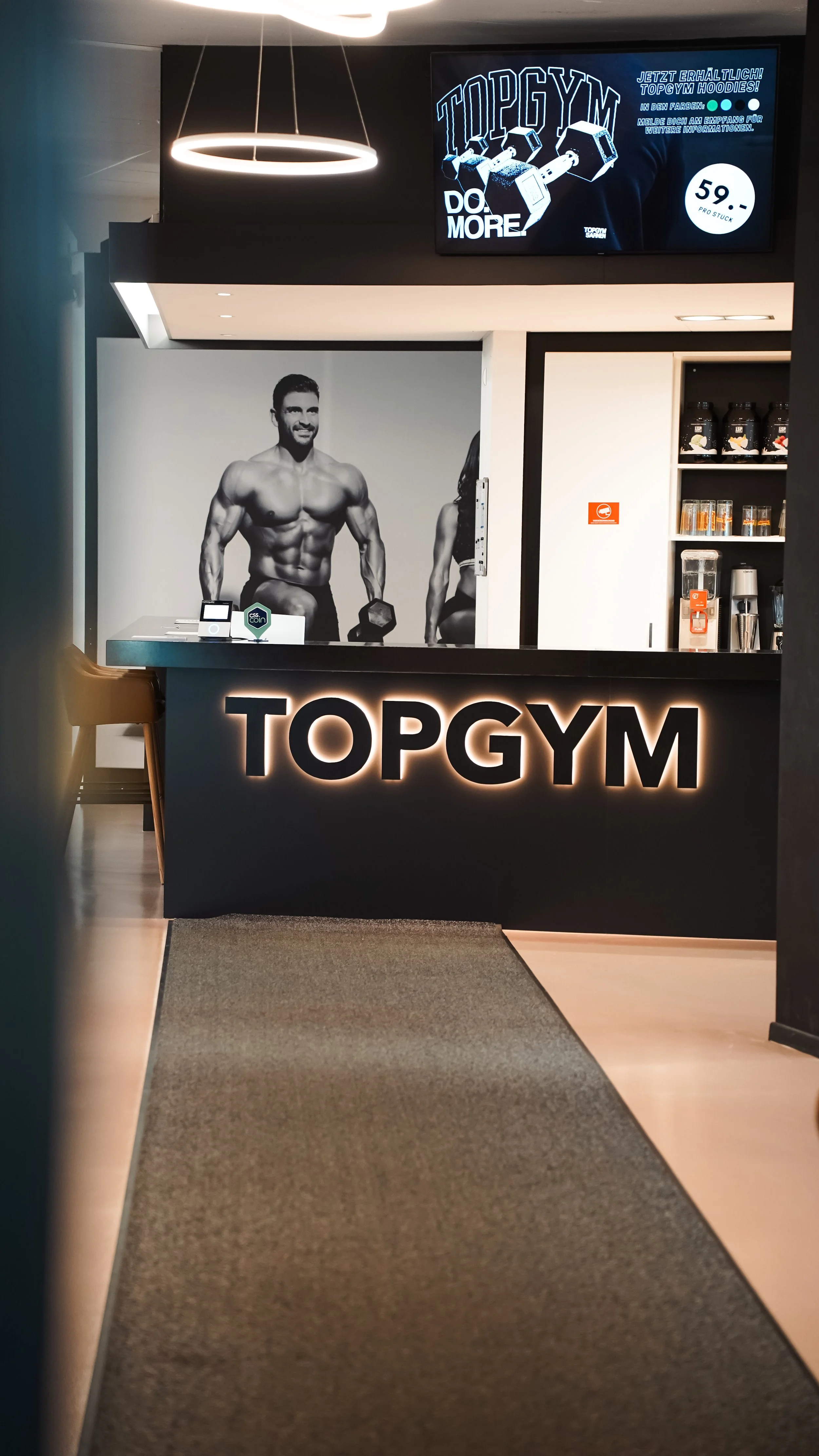 Eingangsbereich eines Fitnessstudios namens 'Topgym' mit beleuchtetem Schild, einer schwarzen Theke mit dem Schriftzug 'TOPGYM' und einem schwarzen und weißen Wandbild eines muskulösen Mannes beim Training.