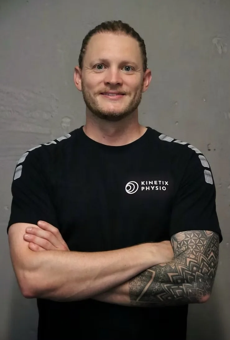 Ein Mann mit kurz gebundenen Haaren und einem Tattoo auf dem linken Arm, der schwarze Sportkleidung mit dem Logo "Kinetix Physio" trägt, steht vor einer grauen Wand mit verschmutzen Stellen und lächelt.