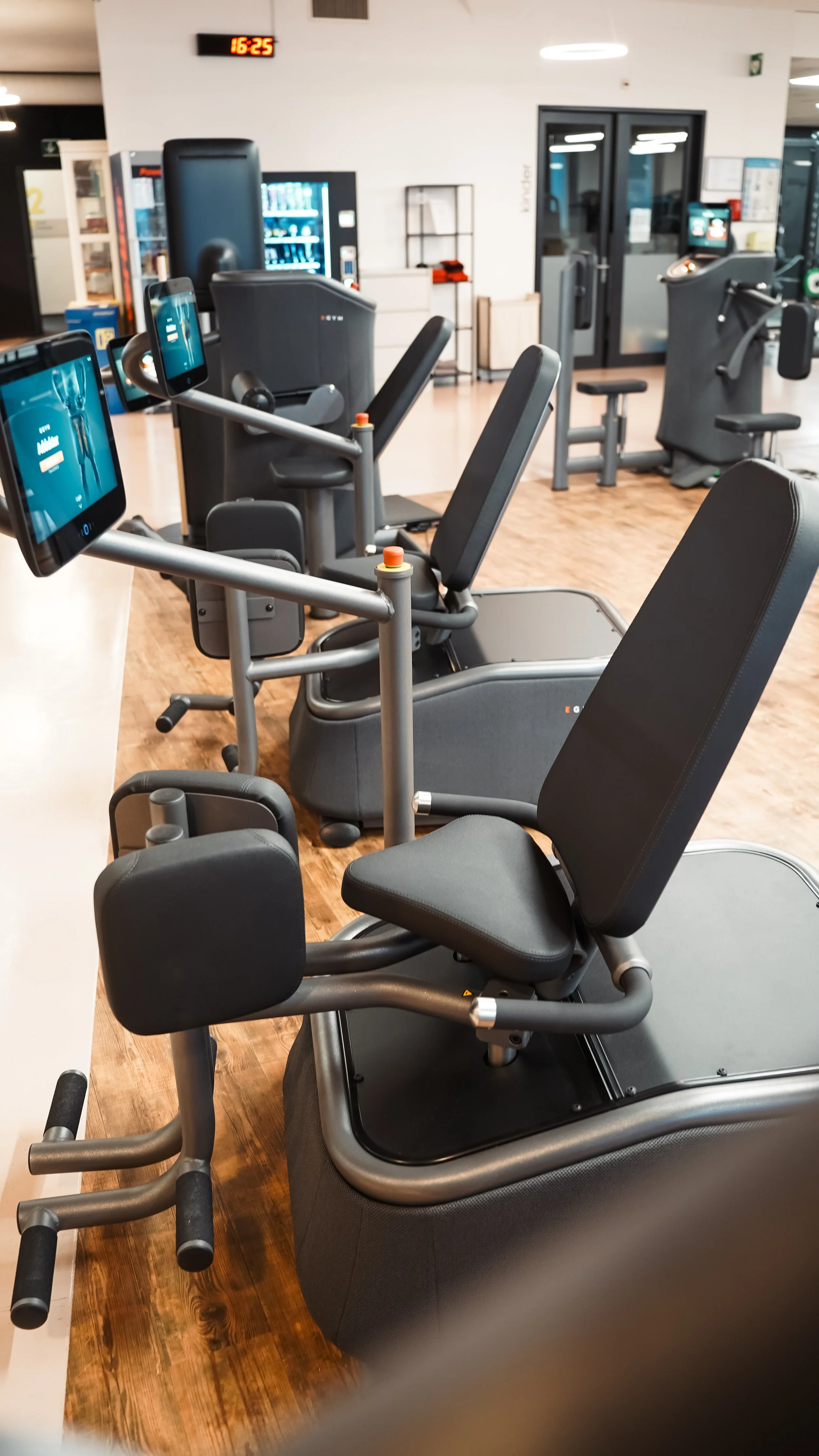 Mehrere Trainingsgeräte im Fitnessstudio, darunter Laufbänder und Sitzmaschinen, mit digitalen Displays, Holzfußboden und einer digitalen Uhr an der Wand.