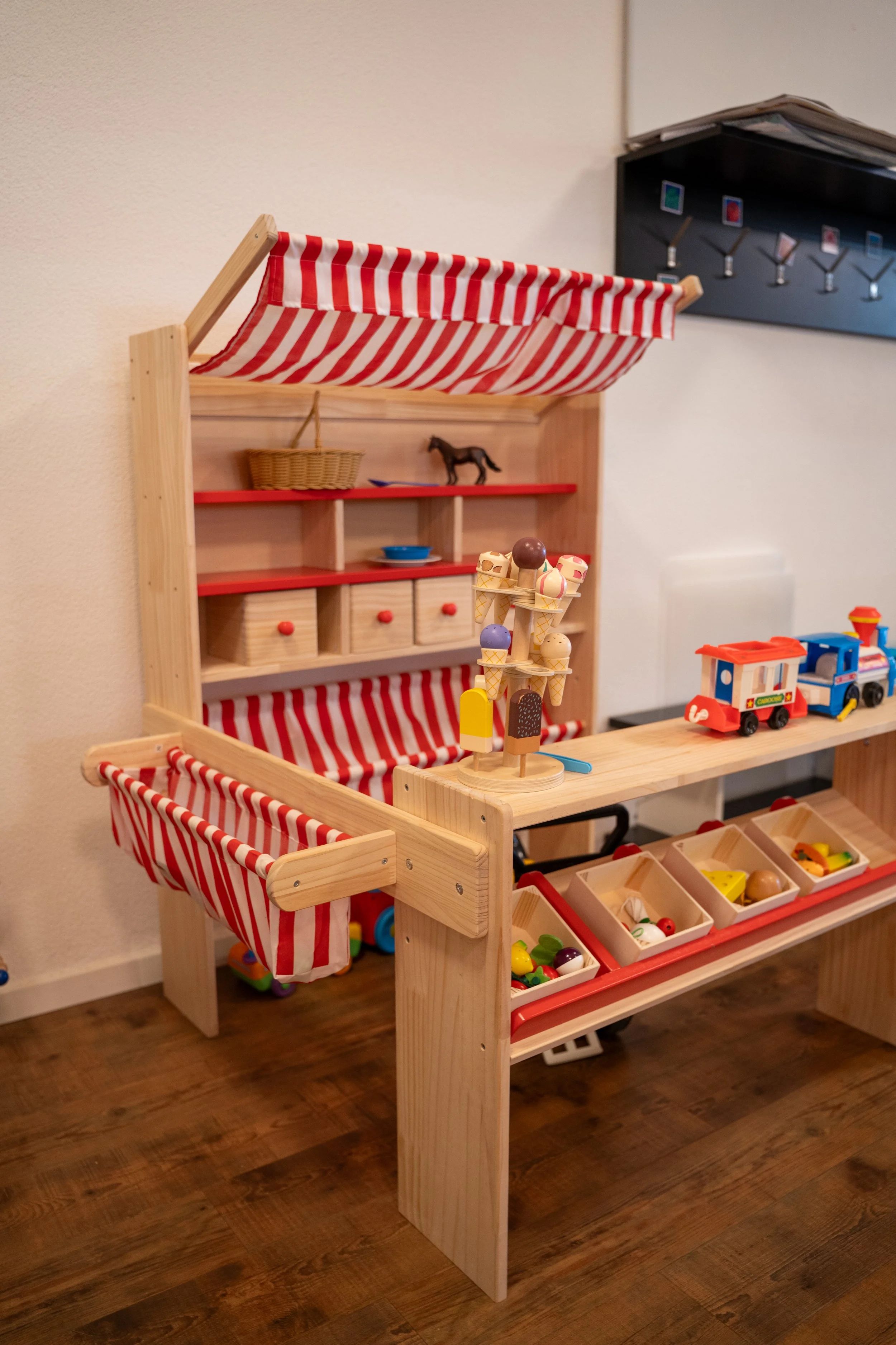 Kleine Holzspielzeugshop mit roten und weißen Streifen Markisen, Spielkörbe, Spielzeugtorten, eine kleine Verkaufsfront mit Spiel Obst und Spiel Lebensmittel, und Spielautos.