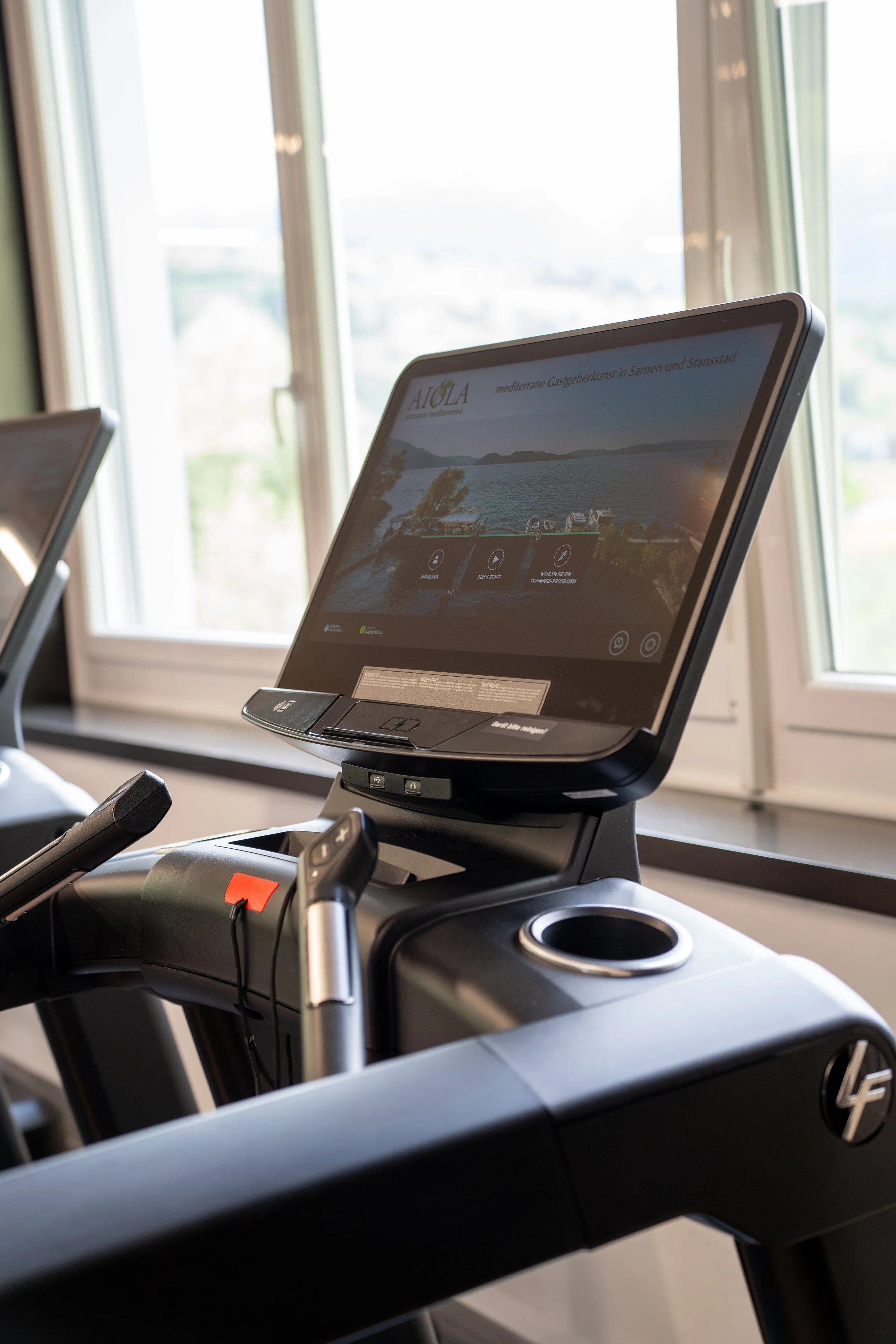 Ein Laufband mit einem Bildschirm, der eine Webseite über eine mediterrane Gegend in Sardinien zeigt, im Fitnessstudio neben einem Fenster.