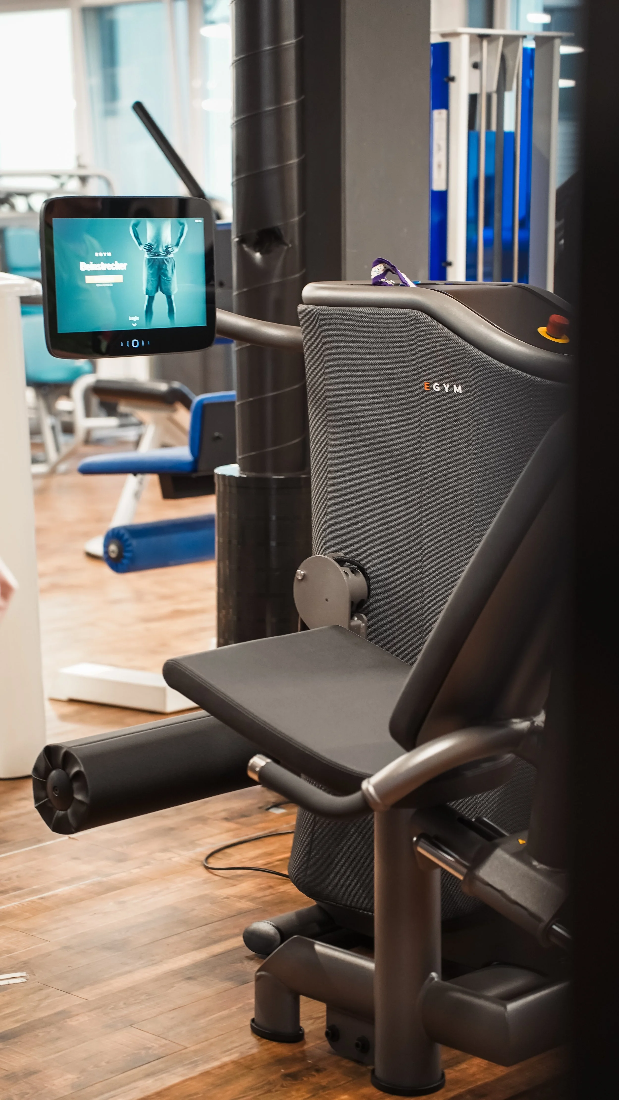 Ein modernes Fitnessgerät im Fitnessstudio mit einem Bildschirm, der einen Menschen beim Training zeigt, und ein Laufband im Hintergrund.