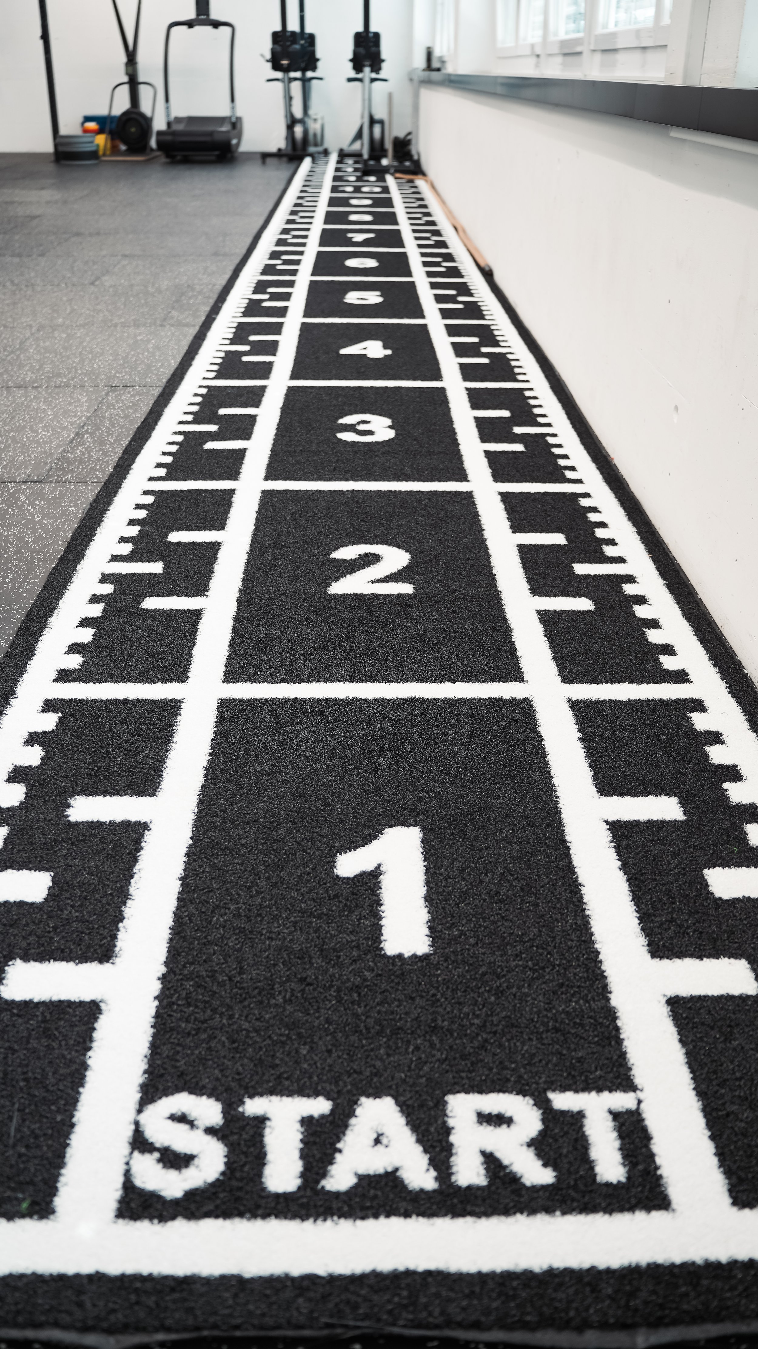 Laufbahn mit Start- und Ziellinien, nummeriert von 1 bis 6, in einem Fitnessstudio mit Trainingsgeräten im Hintergrund.