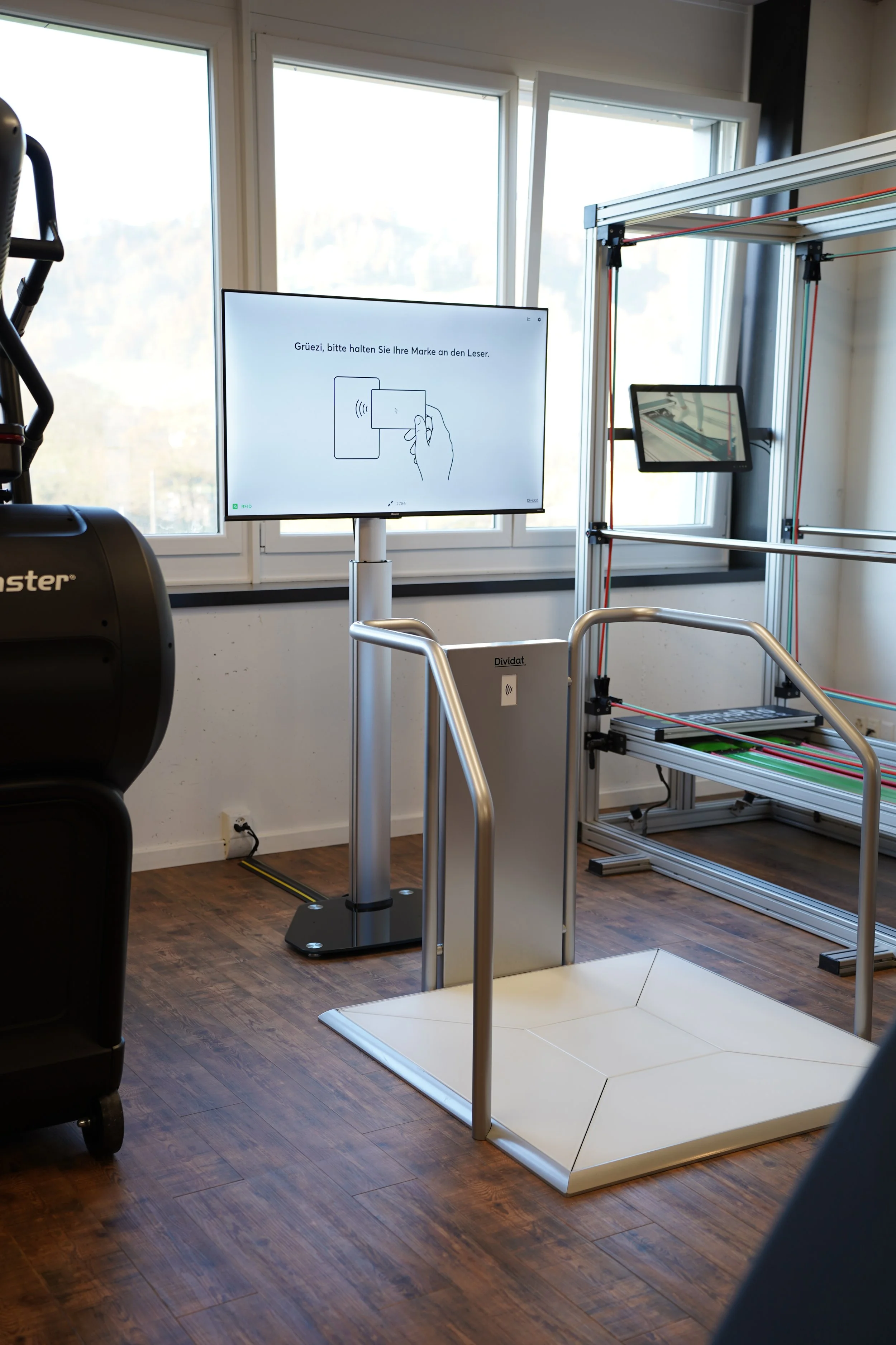 Ein Raum mit einer Waage, einem Bildschirm, einem Fenster und technischen Geräten. Die Waage ist unbemannt, der Bildschirm zeigt eine Anweisung in Deutsch, die das Halten eines RFID-Tags am Leser bittet.