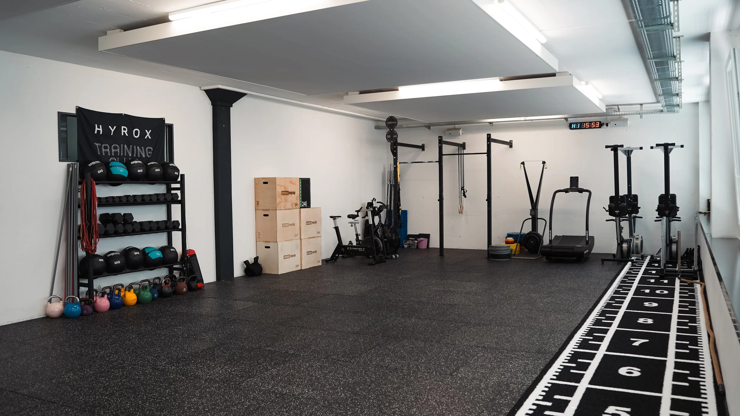 Leerer Fitnessraum mit Gewichten, Kettlebells, Boxen, Spinning-Bikes und Laufbändern auf schwarzem Boden, weißer Wand und großem Fenster. Großes Trainingsmatte mit Messlinien an der Seite.
