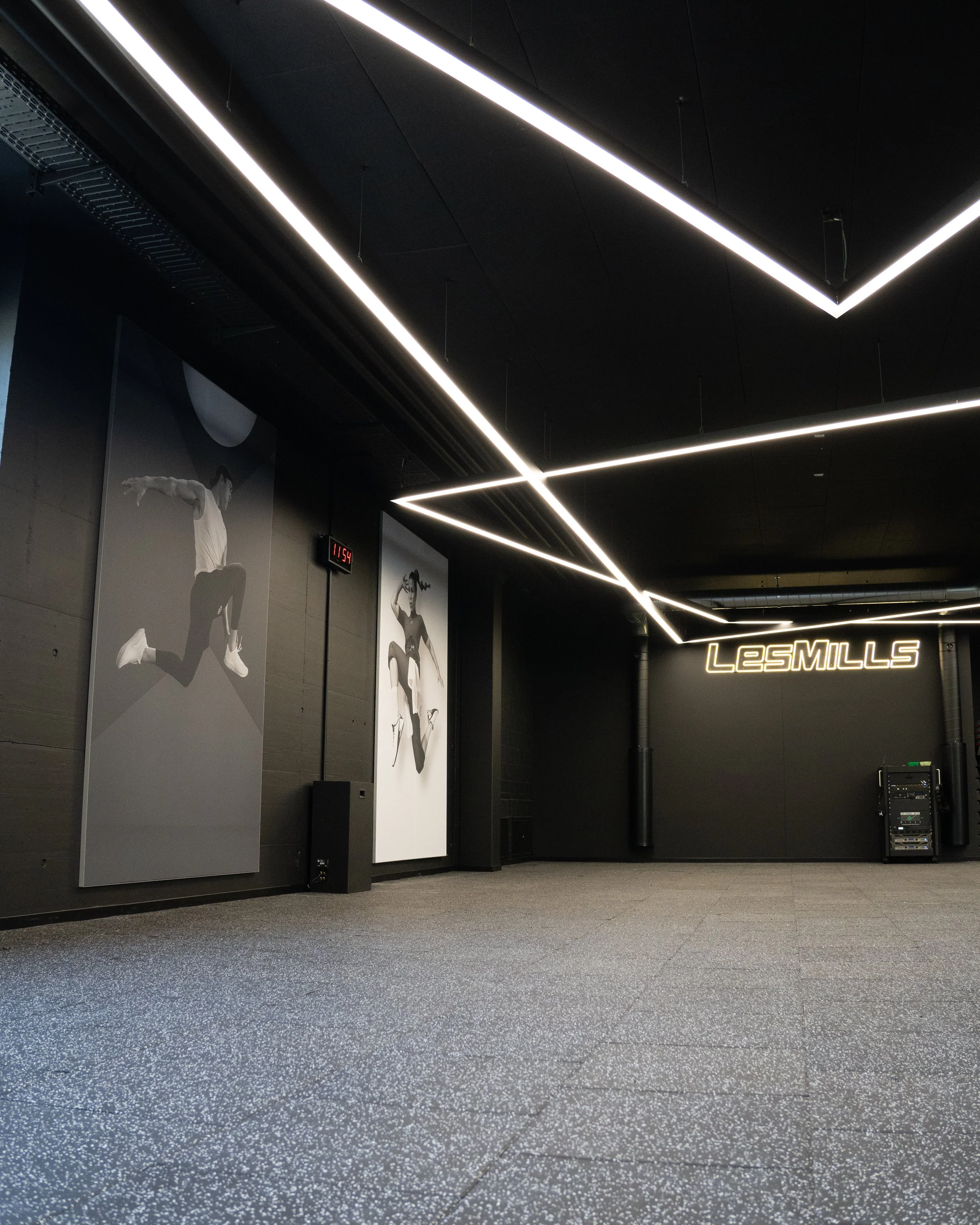 Innenansicht eines modernen Fitnessstudios mit schwarzen Wänden, LED-Lichtleisten an der Decke und großem Leuchtlogo "Les Mills" an der Wand. Es gibt Wandbilder von Sportlern in Aktion.