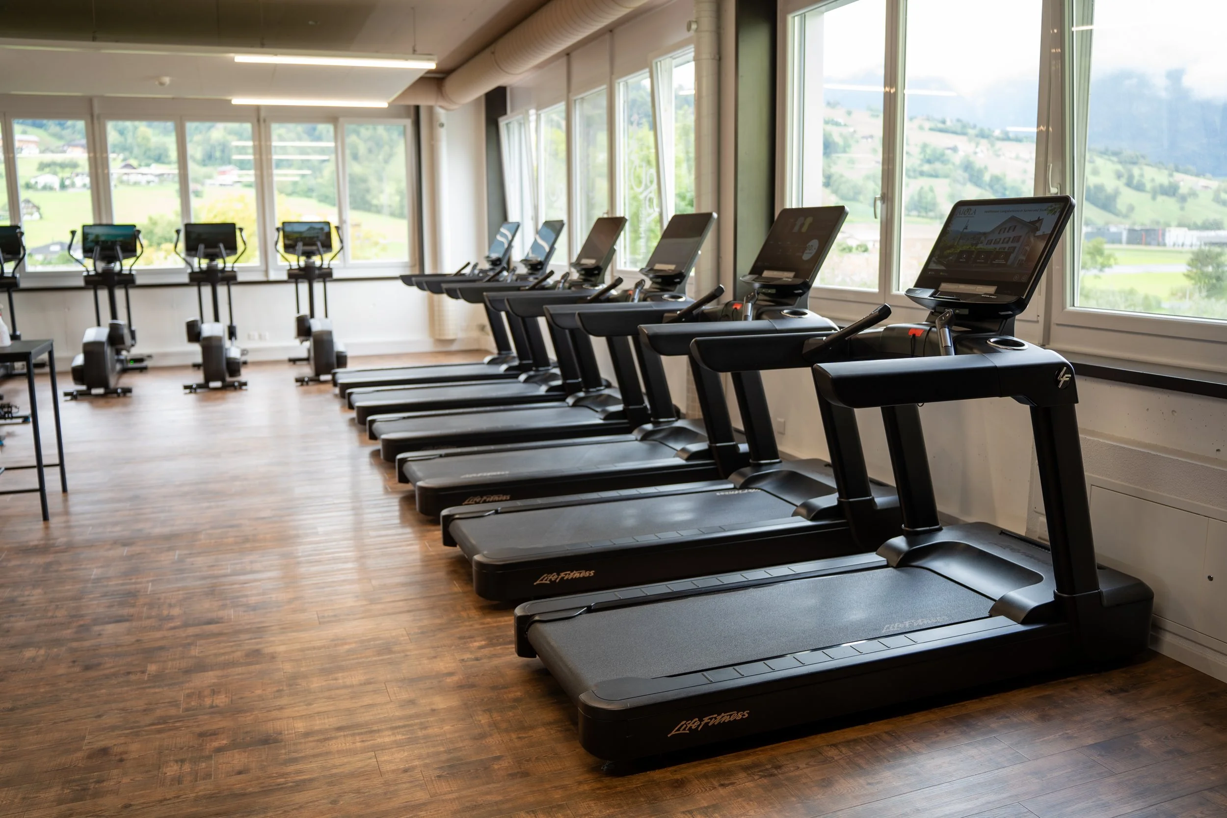 Innenraum eines Fitnessstudios mit mehreren Laufbändern vor großen Fenstern mit Blick auf eine grüne Landschaft.