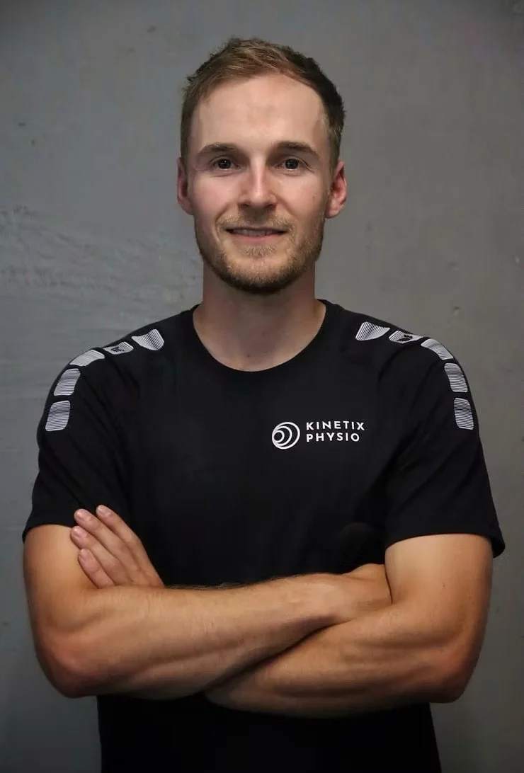 Ein junger Mann mit kurzen braunen Haaren und einem leichten Bart, der ein schwarzes T-Shirt mit weißen Akzenten und dem Logo 'Kinetix Physio' trägt, steht vor einer grauen Wand und lächelt. Er hat die Arme verschränkt.