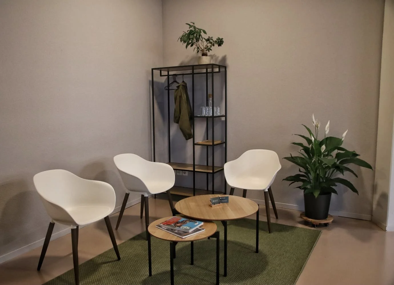 Warteraum mit drei weißen Stühlen um einen kleinen runden Tisch, einem größeren runden Tisch, einem grünen Teppich und Pflanzen in Töpfen, einer schwarzen Metalleinrichtung im Hintergrund und neutralen Wänden.
