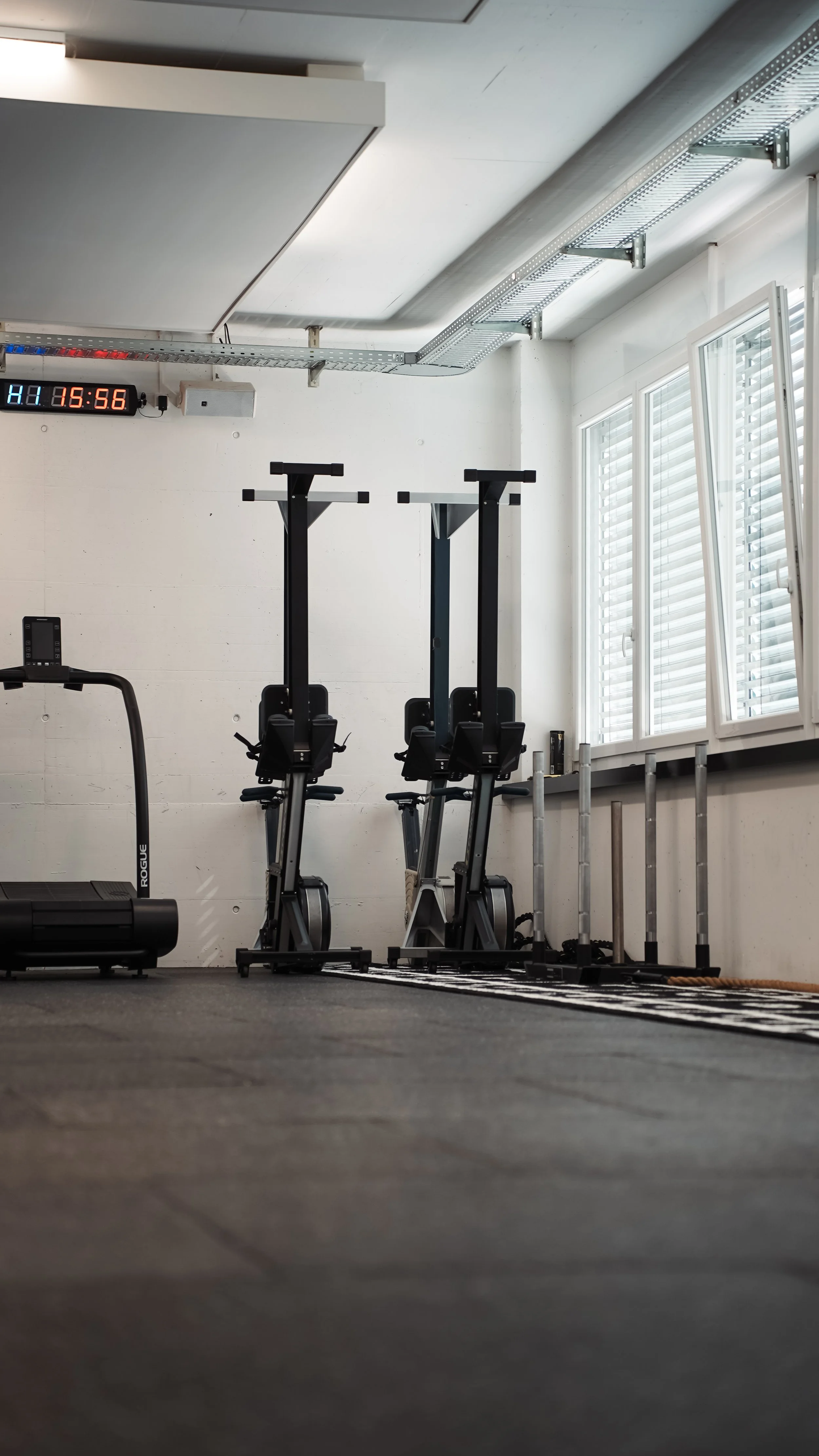 Leerer Fitnessraum mit zwei Steppern, einem Laufband, einem vertikalen Timer an der Wand, einer Fensterfront mit offenen Jalousien und einer schwarzen Gymnastikmatte auf dem Boden.