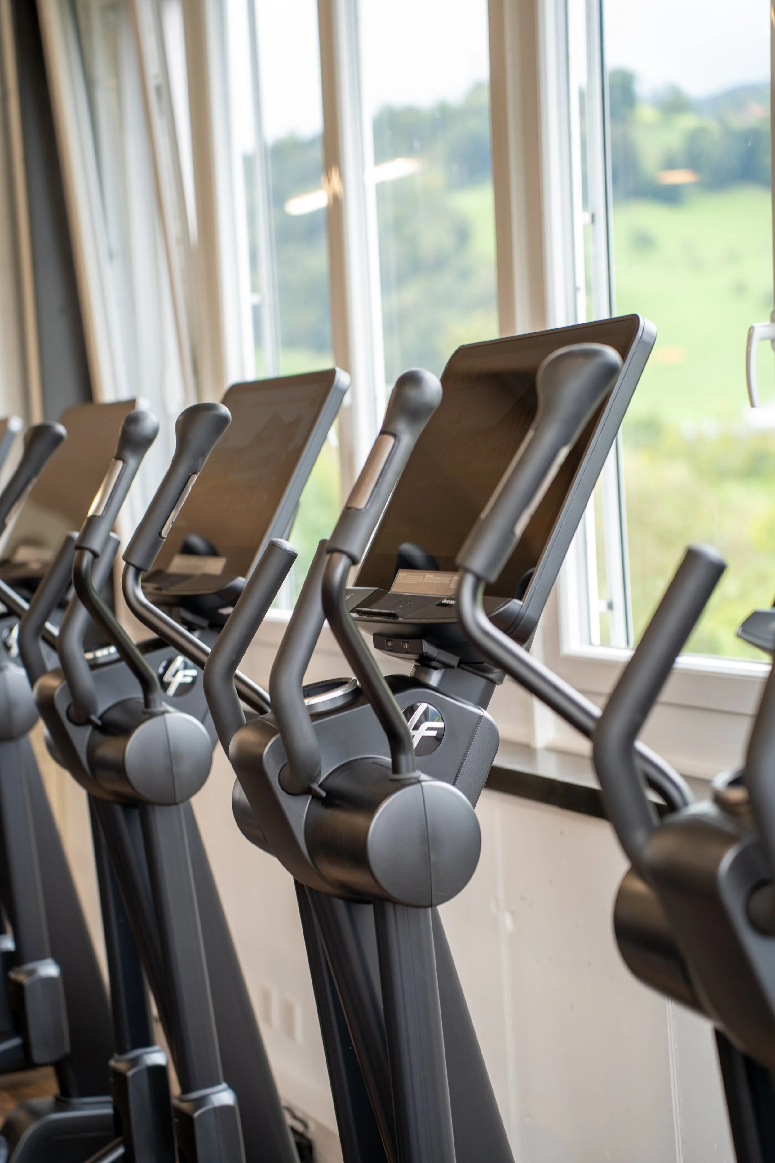 Mehrere elliptische Fitnessgeräte mit Bildschirmen, die in einem Fitnessstudio vor einem Fenster stehen, das eine grüne Landschaft zeigt.