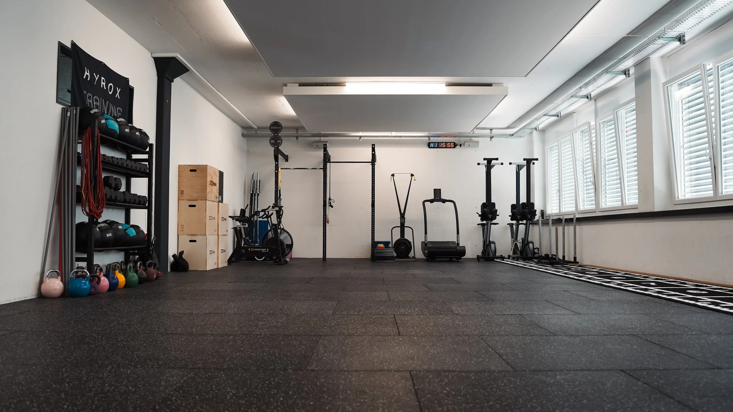 Leeres Fitnessstudio mit Hanteln, Kettlebells und Trainingsgeräten auf schwarzem Gummiboden, große Fenster an der rechten Wand