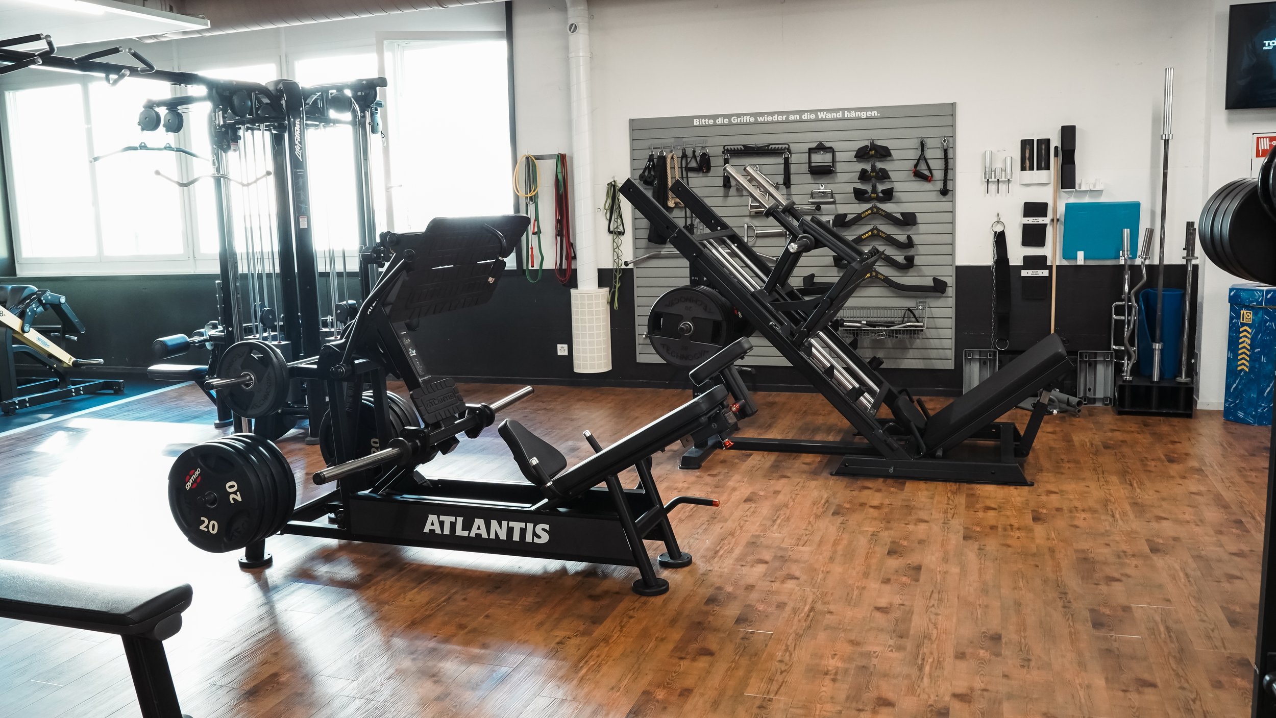 Innenaufnahme eines Fitnessstudios mit Krafttrainingsgeräten, Hanteln, Handgriffen und Wandhalterungen für Zubehör, Holzfußboden und Fenster