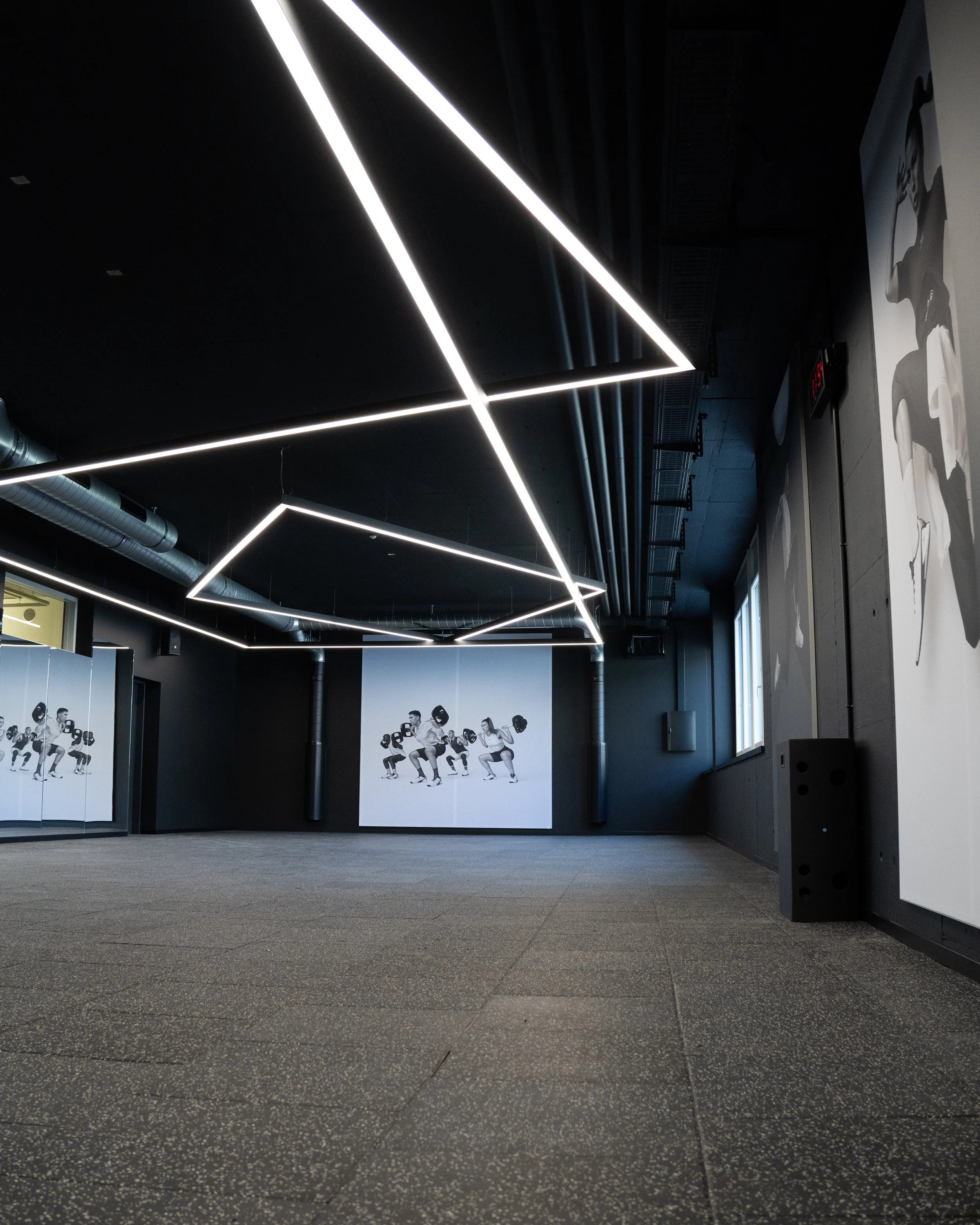 Ein moderner Fitnessraum mit schwarzen Wänden, geometrischen Lichtinstallationen an der Decke und Wandbildern von Menschen, die Gewichte heben.