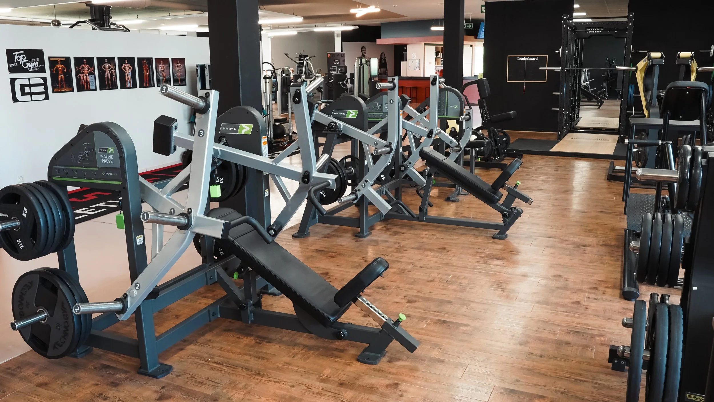 Fitnessstudio mit verschiedenen Trainingsgeräten, Hanteln und Kraftmaschinen, Holzfußboden, Wandbilder von Muskelmuskeln.