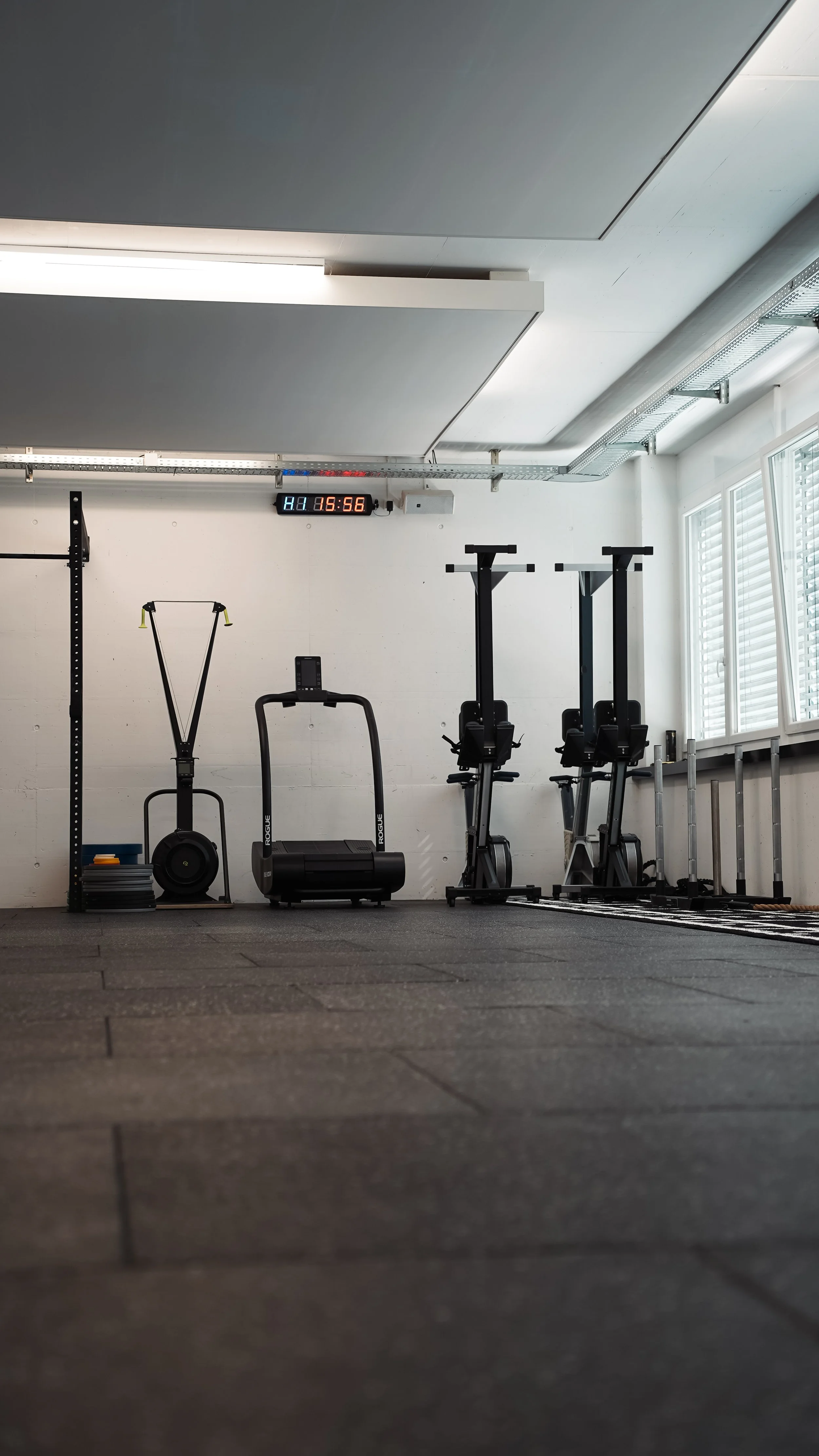 Leerer Fitnessraum mit Laufband, Rudergerät, Fahrradergometern und anderen Trainingsgeräten, Sonnenlicht durch Fenster, digitale Zeituhr an der Wand.